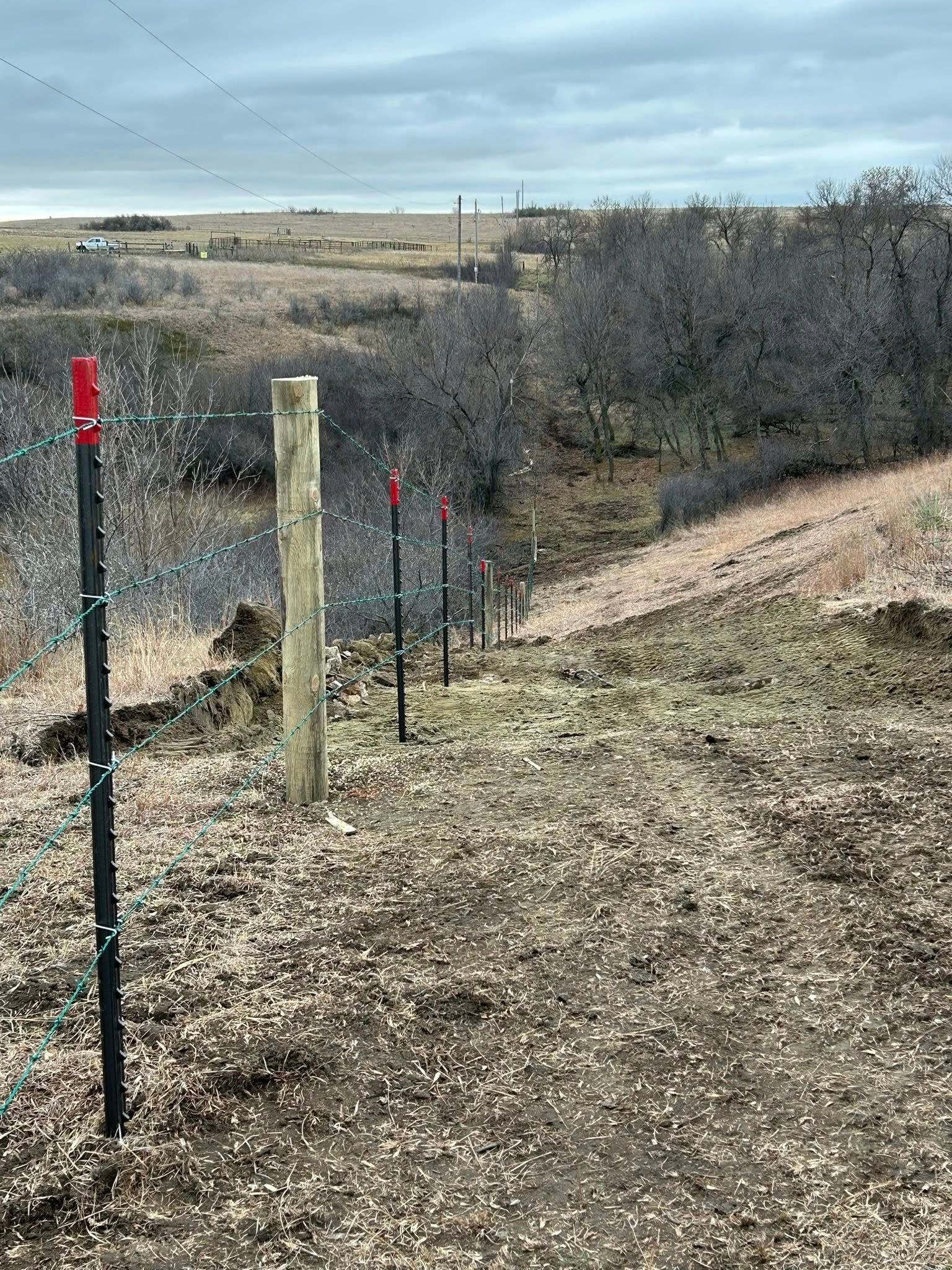 A fence with black posts and wire leads down a hillside. The posts are marked with red tips.