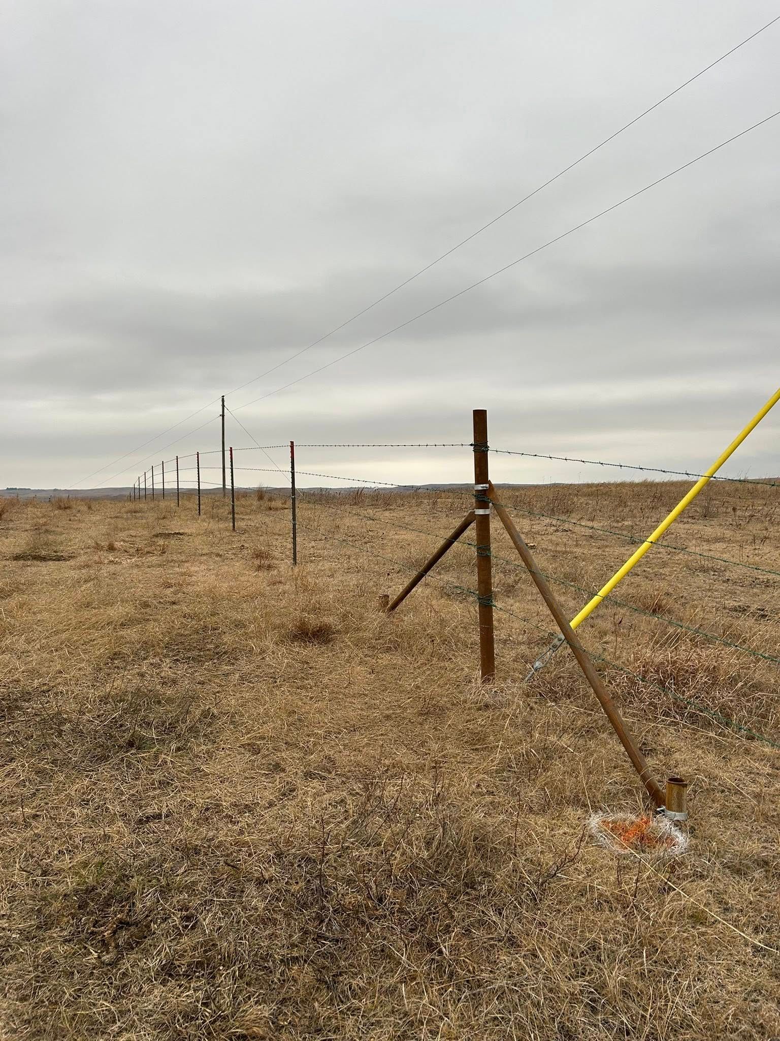 Brown wooden posts and white poles in a field, holding up lines in a row against a cloudy sky.