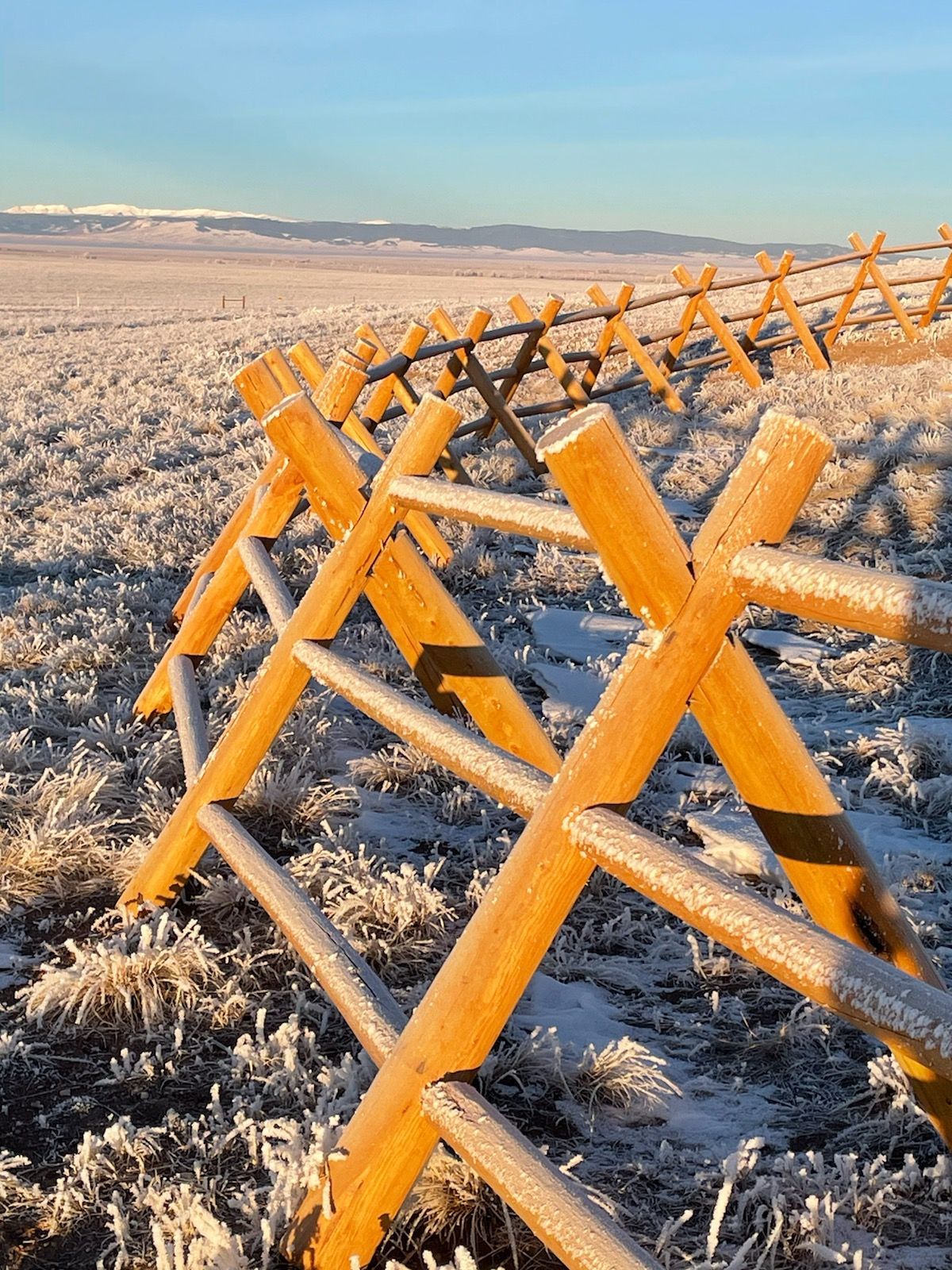 Wooden split-rail fence in a frost-covered field with mountains in the background under a blue sky.