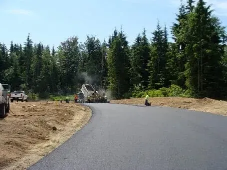 Curved freshly paved road beside a forest, with construction equipment and workers in the distance