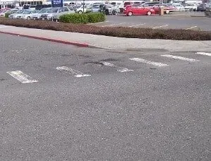 Parking lot roadway with faded white crosswalk markings and a curbside median with shrubs.
