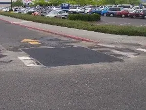 Damaged asphalt patch across a parking lot entrance near parked cars and a curb