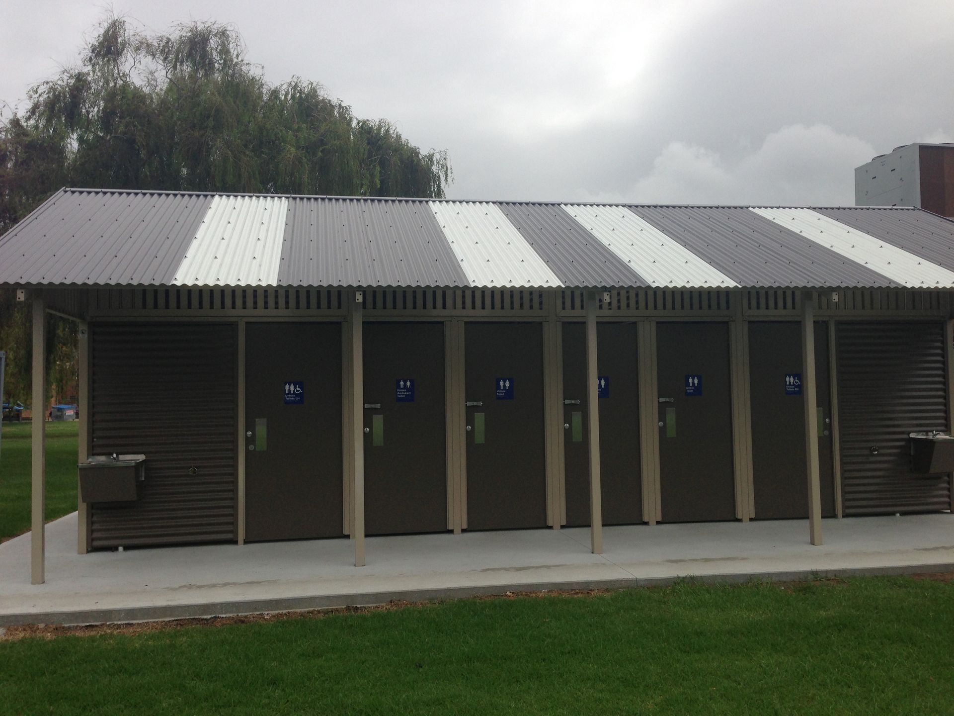 Public restroom with corrugated metal roof and doors, grey/white striped roof, grass in front.