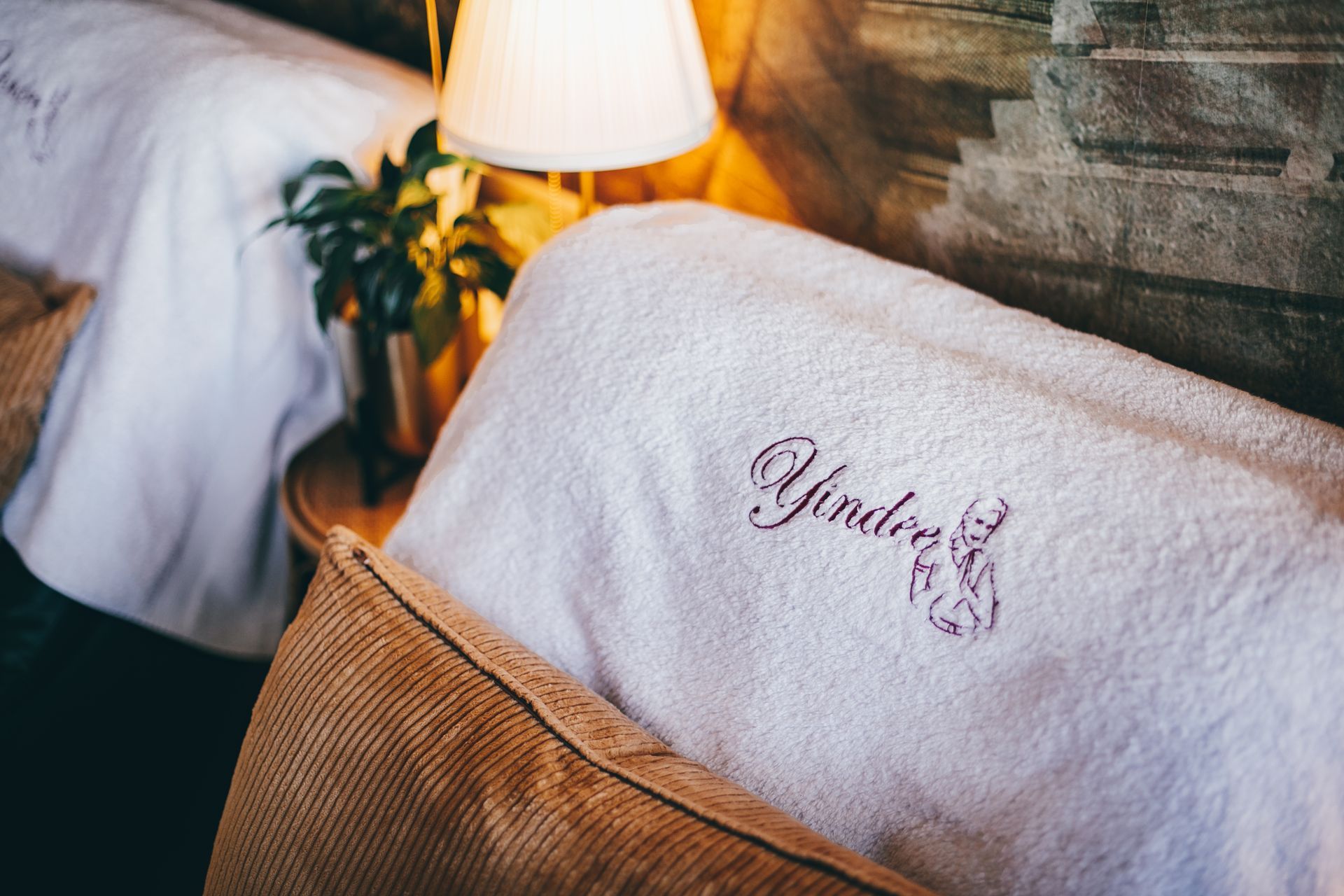 Two white towels with embroidered script on beds, bedside lamp, and a plant.