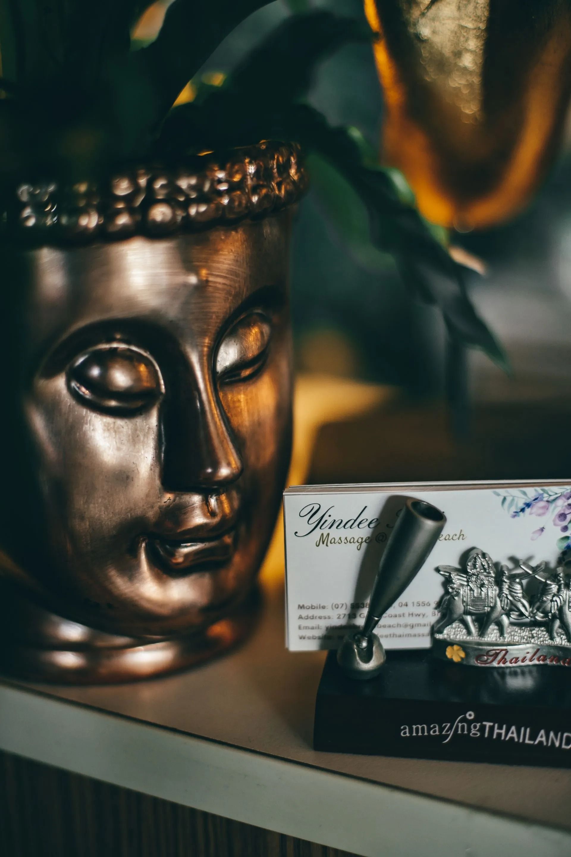 Bronze Buddha Head, Pen Holder, and Business Card on a Shelf, Near a Plant — Traditional Massage Services In Broadbeach, QLD