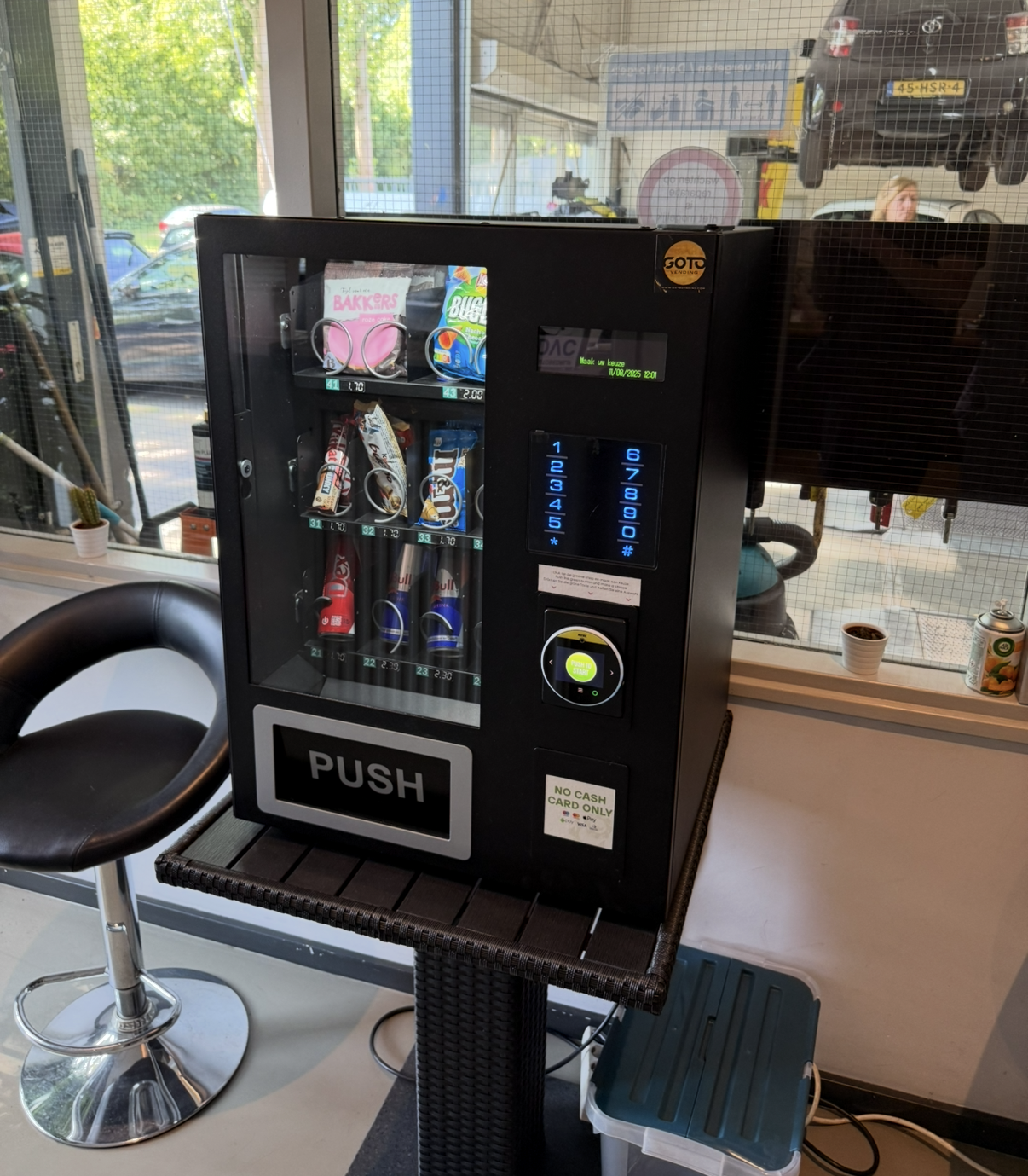 Een zwarte automaat met diverse snacks, die op een sokkel in een winkel met een raam en een barkruk staat.