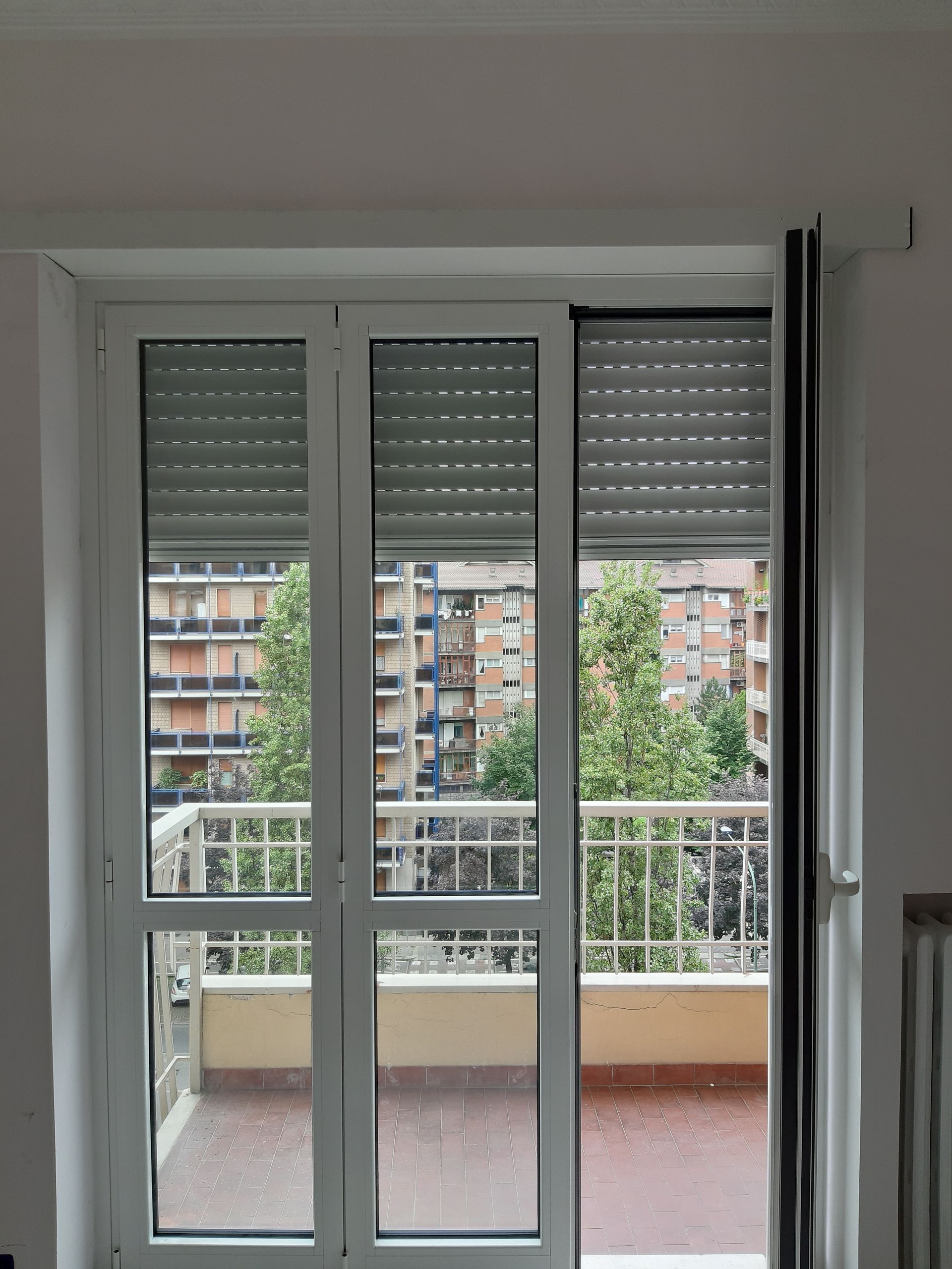 Porta-finestra bianca con persiane avvolgibili, che si affaccia su un balcone con ringhiera e sugli edifici cittadini.