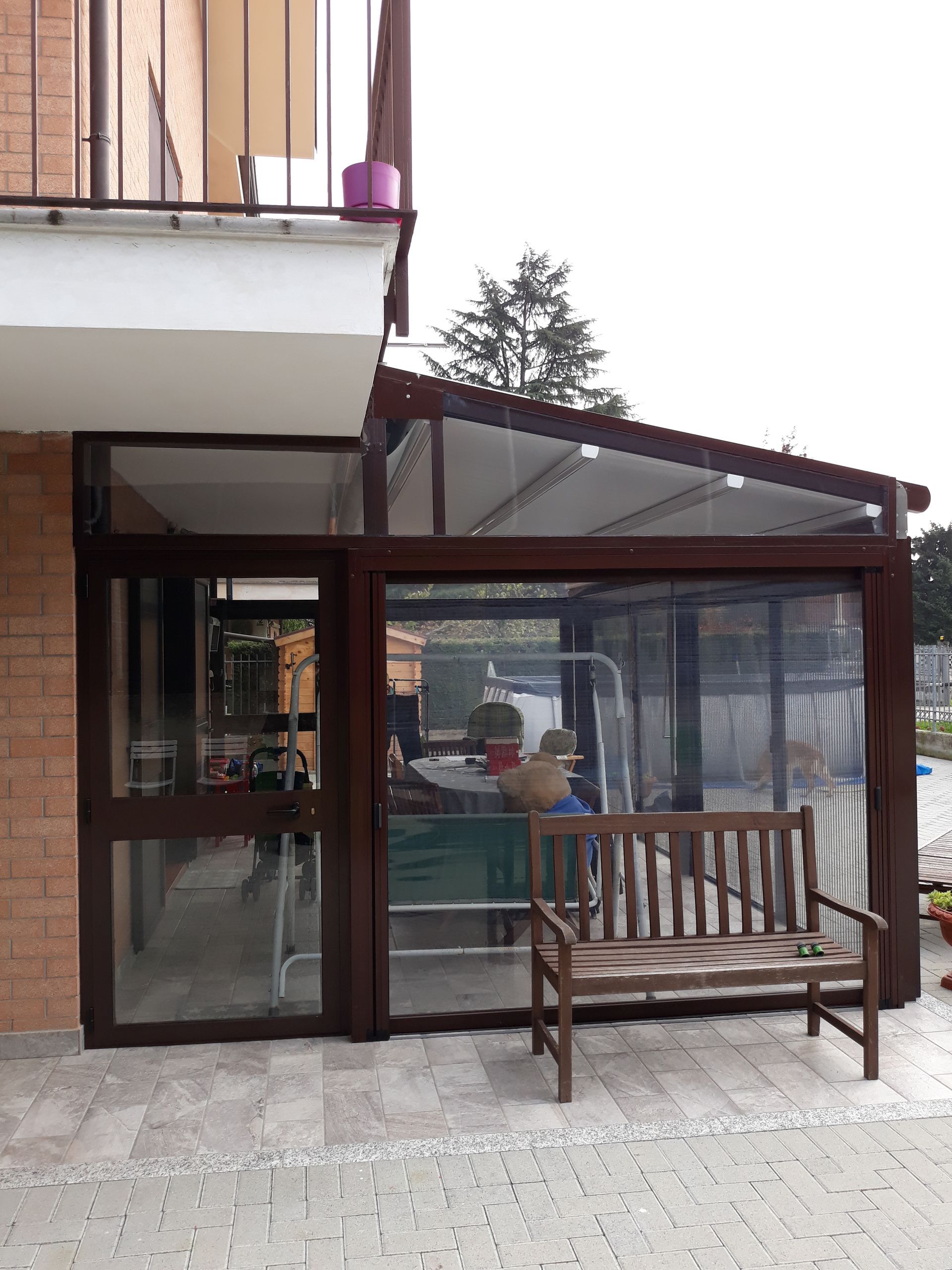 Veranda chiusa con finiture marroni e una panca in legno; annessa a un edificio bianco con balcone.