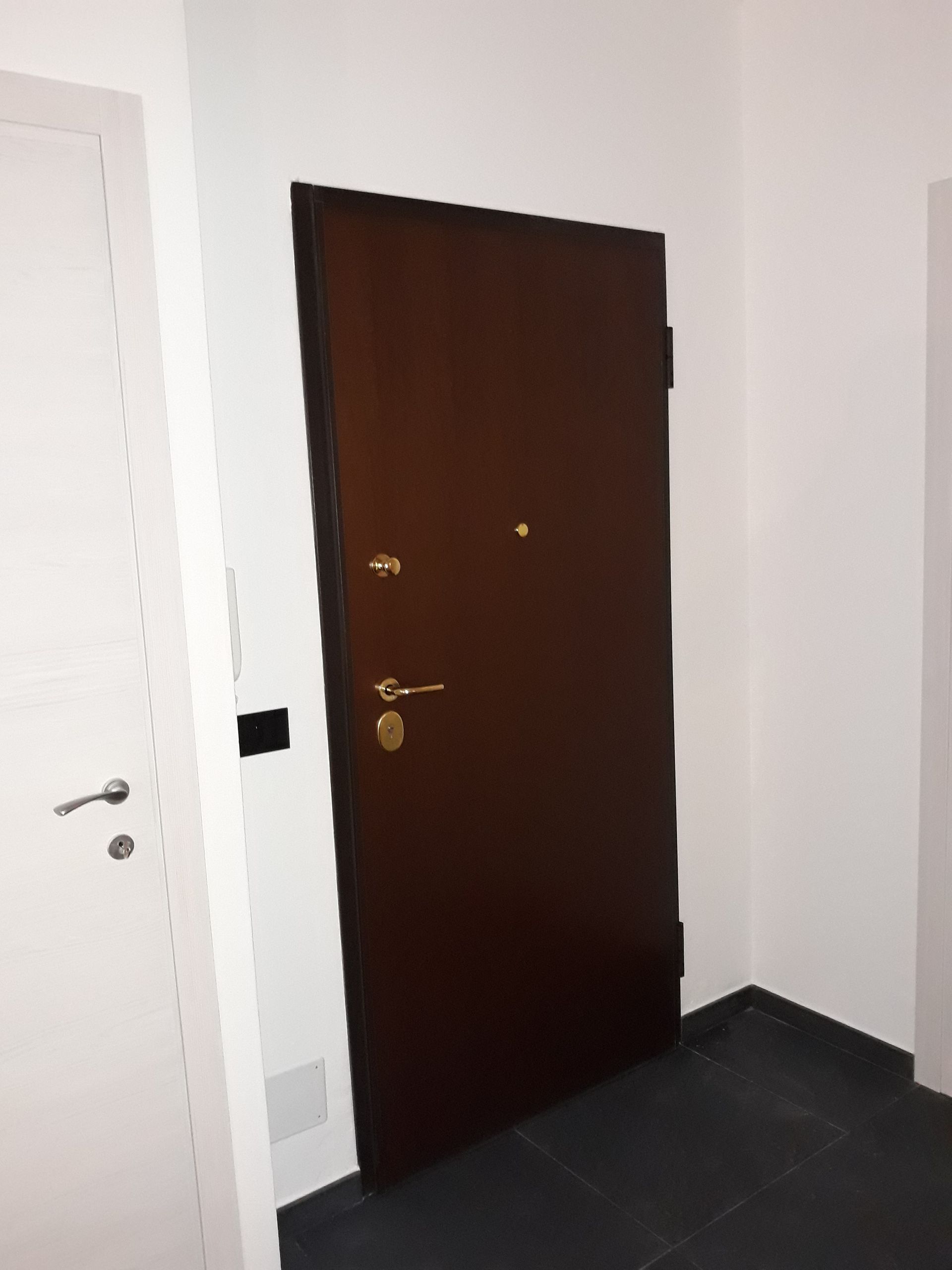 Porta marrone in una stanza bianca con una porta bianca sulla sinistra e un pavimento piastrellato nero.