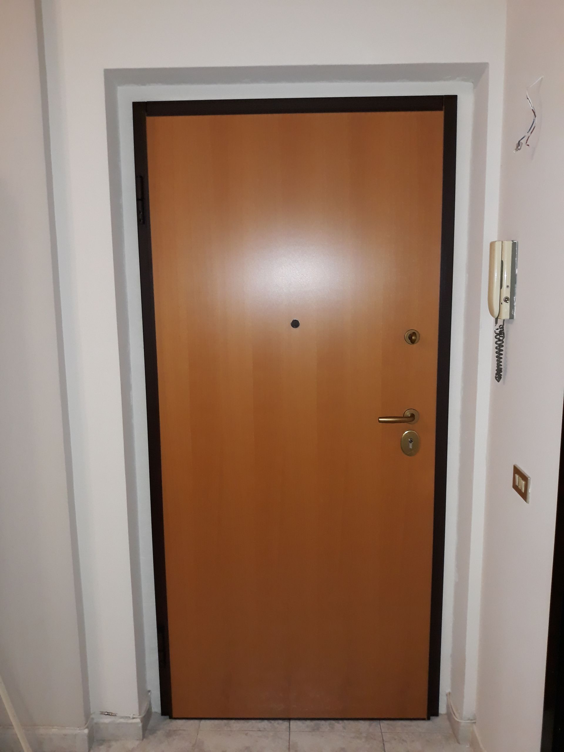 Porta in legno con finiture e ferramenta nere su un ingresso bianco, con campanello e citofono sulla parete.