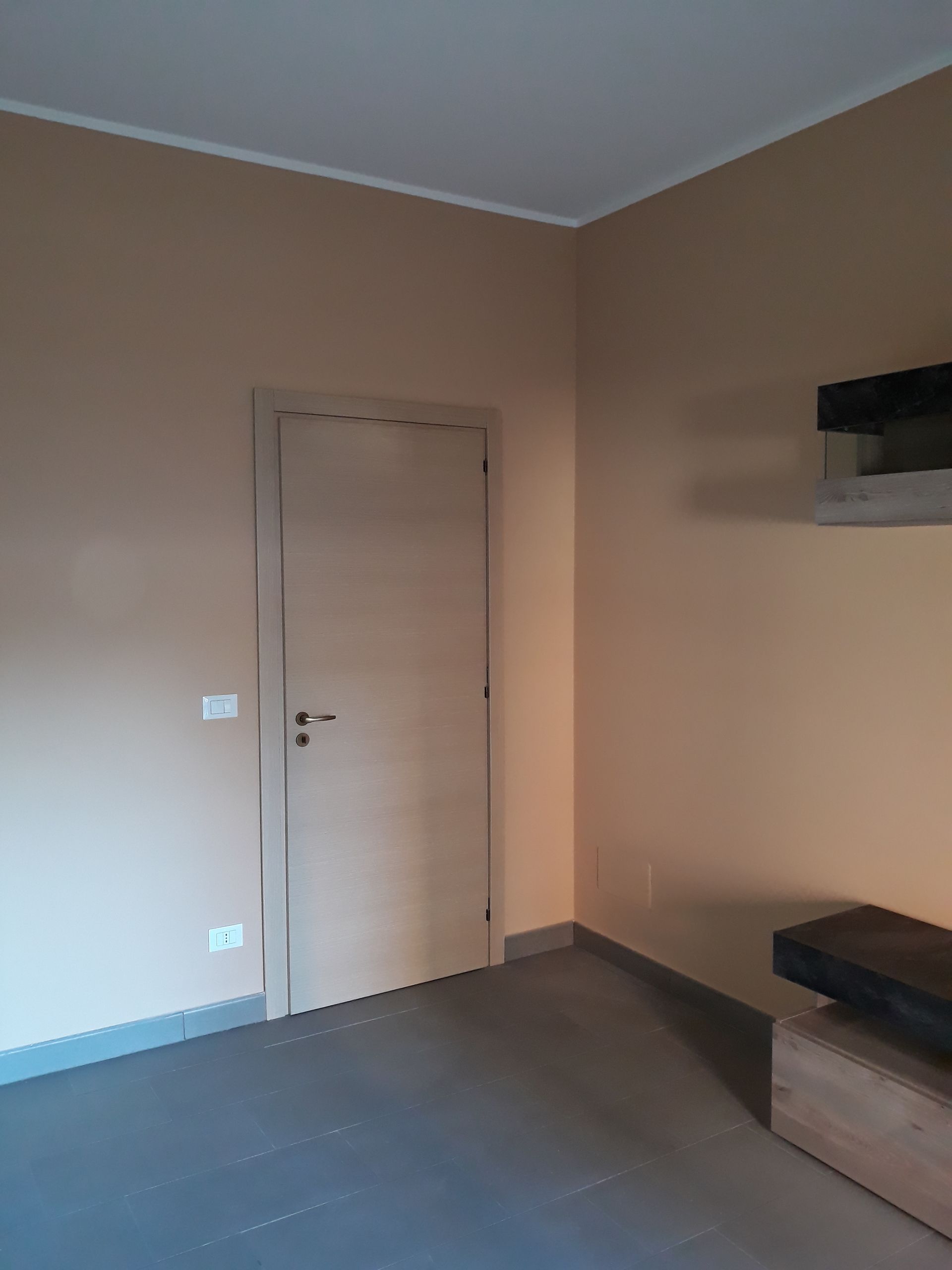 Stanza con pareti beige, porta chiusa, pavimento grigio e un mobile scuro. Il soffitto è decorato con profili bianchi.