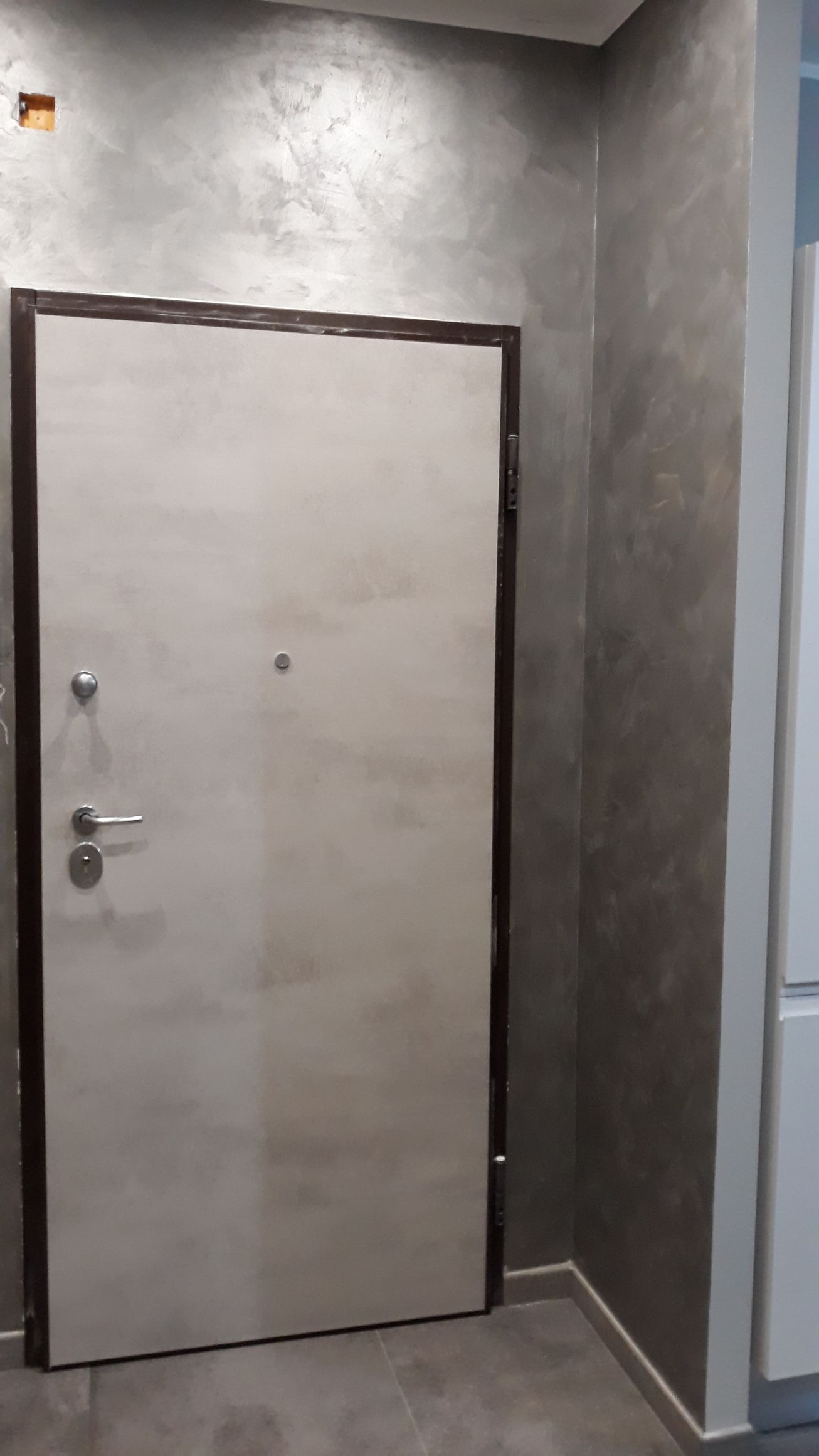 Una porta d'ingresso beige con finiture scure, inserita in un corridoio con pareti grigie testurizzate.