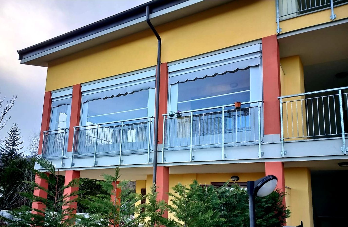 Edificio giallo con balconi in vetro trasparente, finiture arancioni e tende da sole.