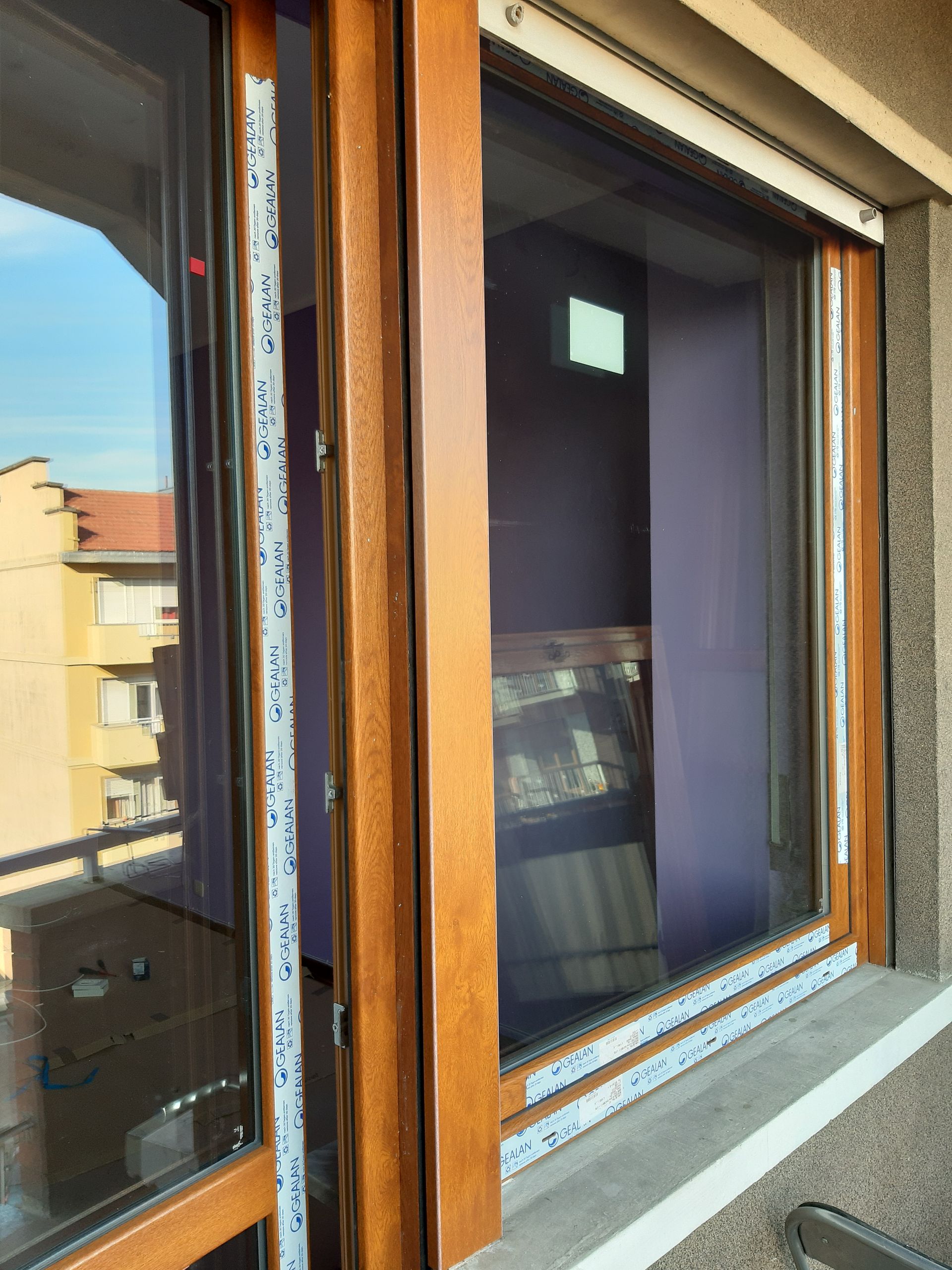 Finestra con cornice marrone, parzialmente aperta, con vista sugli edifici. All'interno è visibile una tenda viola.