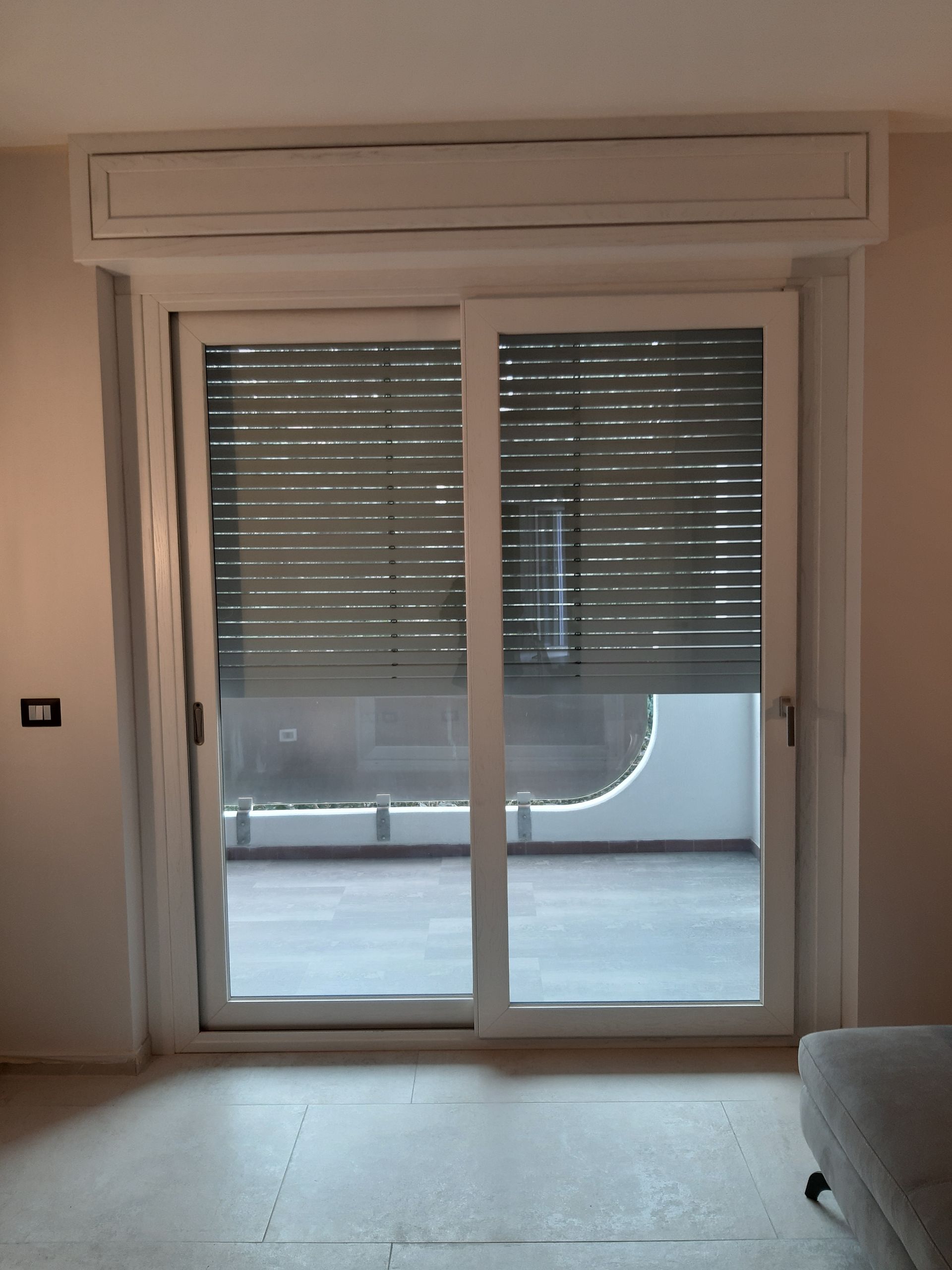 Porta scorrevole in vetro con persiane chiuse e vista su un balcone. Inquadratura interna, colori neutri.