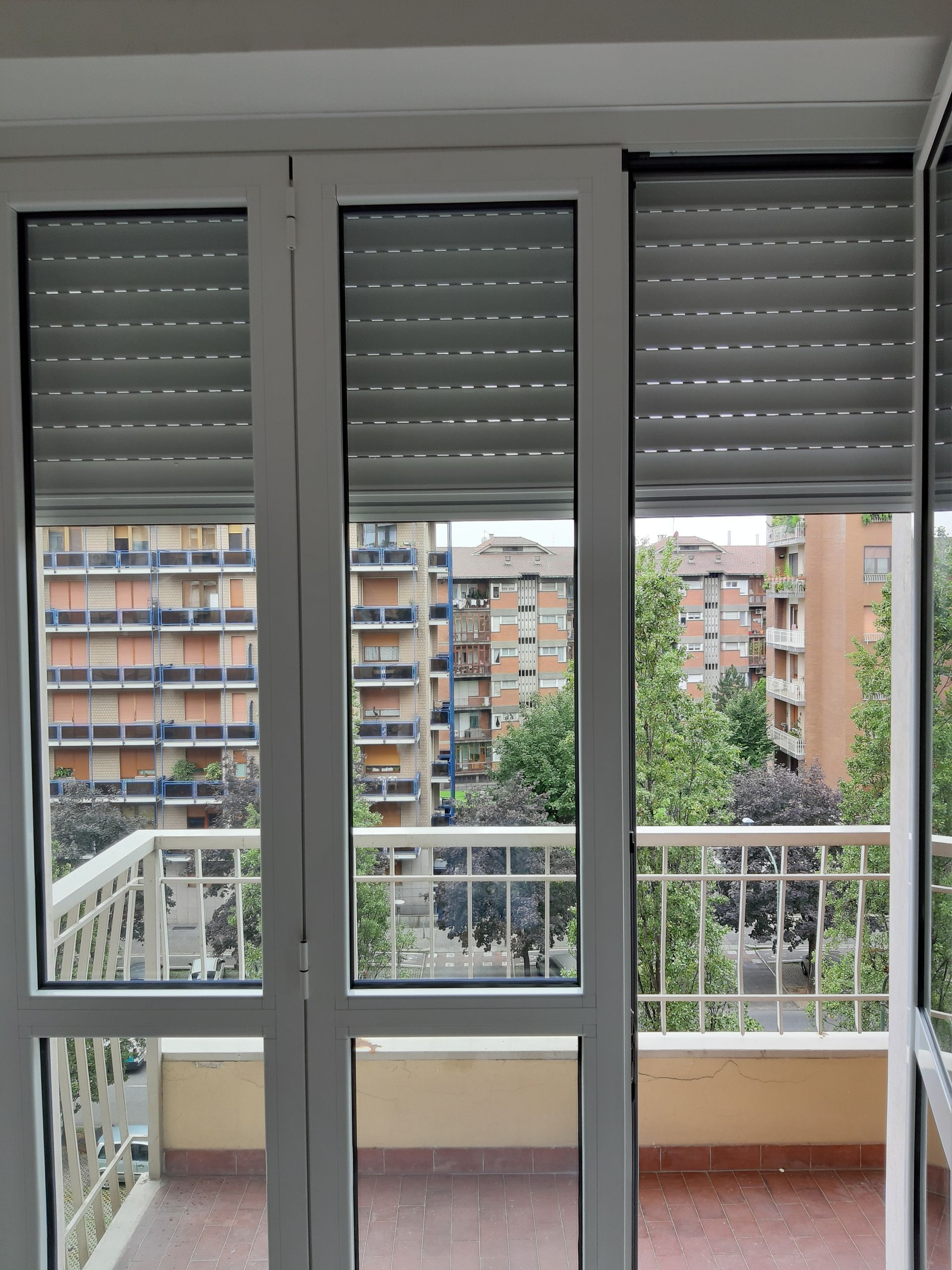 Vista dal balcone con finestre con cornice bianca e persiane grigie chiuse, con vista su edifici e alberi.