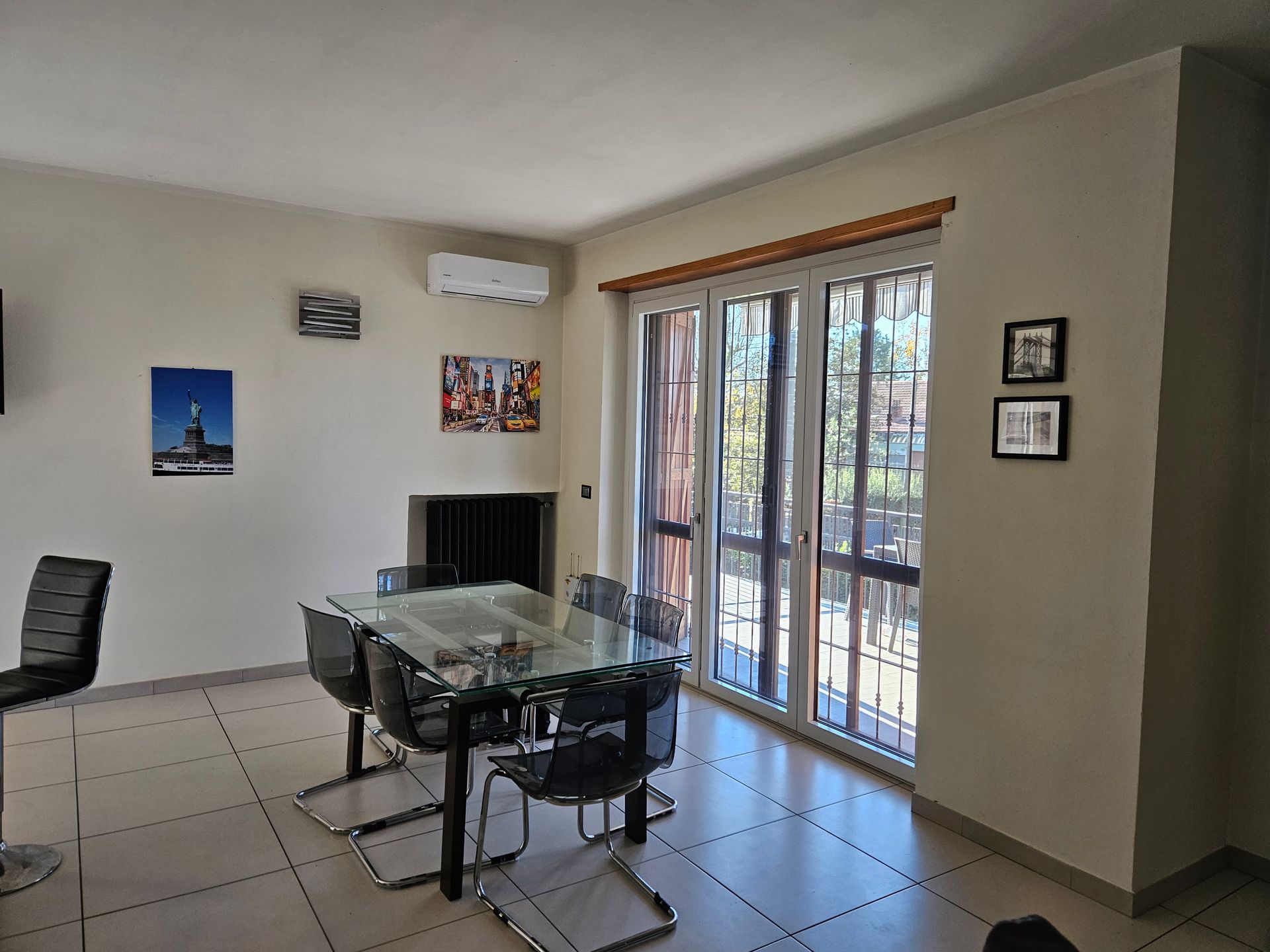 Sala da pranzo con tavolo in vetro e sedie nere, vicino a una porta scorrevole. Sono visibili anche quadri alle pareti e aria condizionata.
