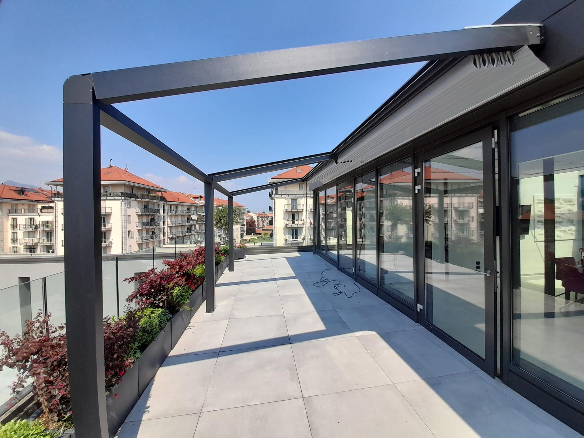 Moderno patio sul tetto con pergolato, porte in vetro e piante rosse sullo sfondo del paesaggio urbano.