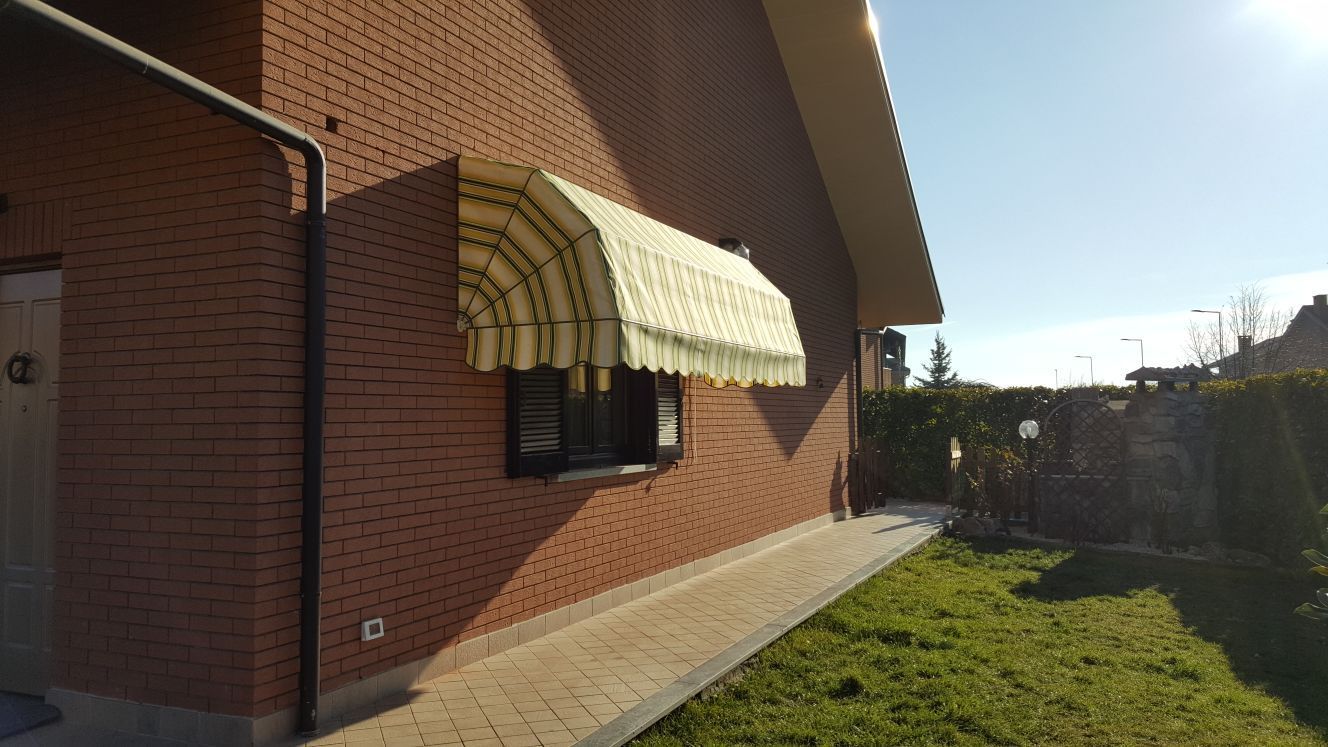 Esterno di una casa in mattoni con una tenda da sole sopra una finestra, un cortile erboso e un cielo soleggiato.