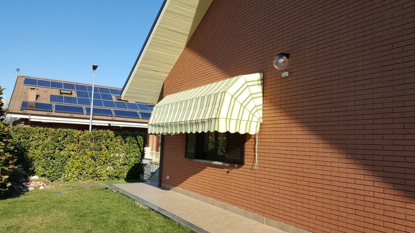 Casa in mattoni con una tenda a strisce sopra una finestra, giornata di sole. Pannelli solari sul tetto sullo sfondo.