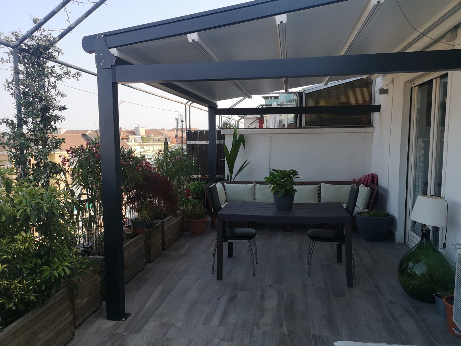 Patio esterno con pergolato nero, tavolo, sedie e sedute integrate. Piante e vista sulla città visibili.