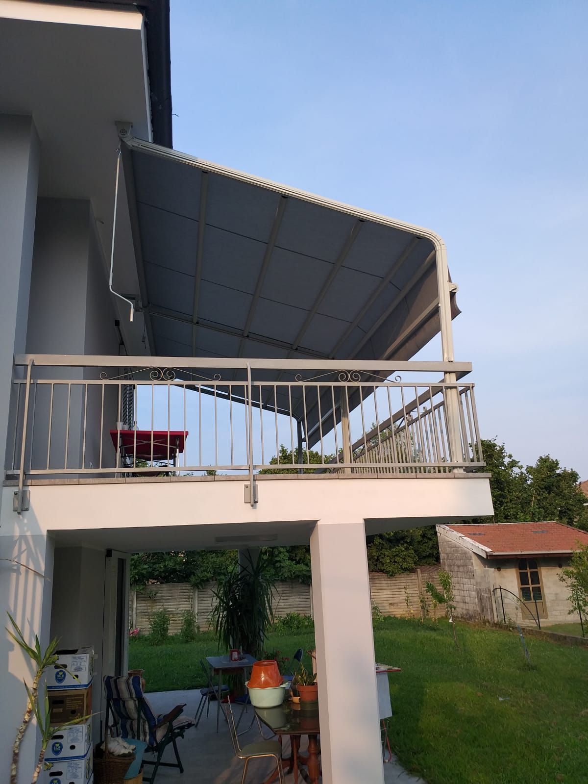 Balcone con ringhiera in metallo e tetto grigio ondulato, con vista su un cortile con capanno.