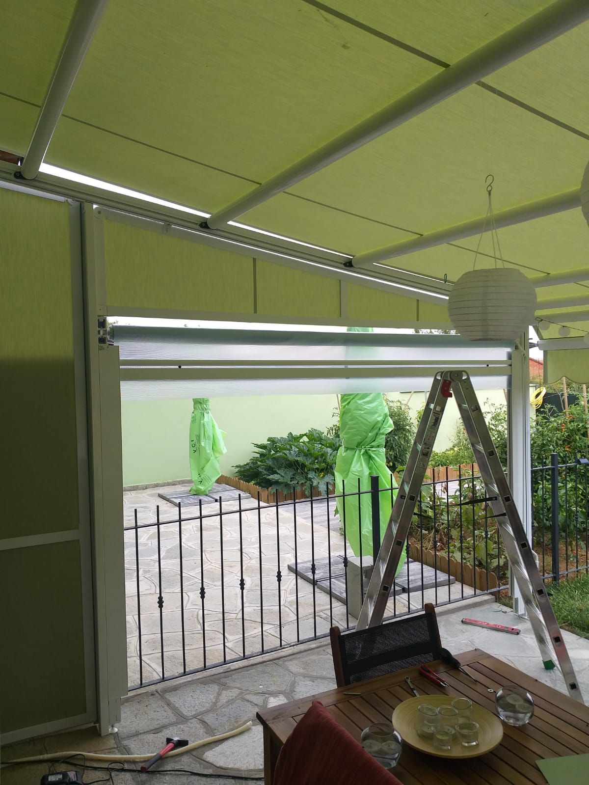 Tenda verde sopra il patio con cancello metallico aperto, vista sul cortile, sugli alberi e sugli ombrelloni coperti.