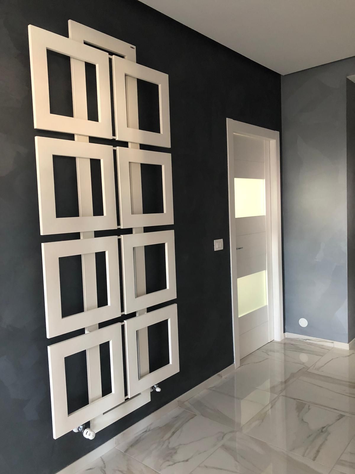 Radiatore geometrico bianco su una parete grigio scuro, porta bianca e pavimento in marmo.