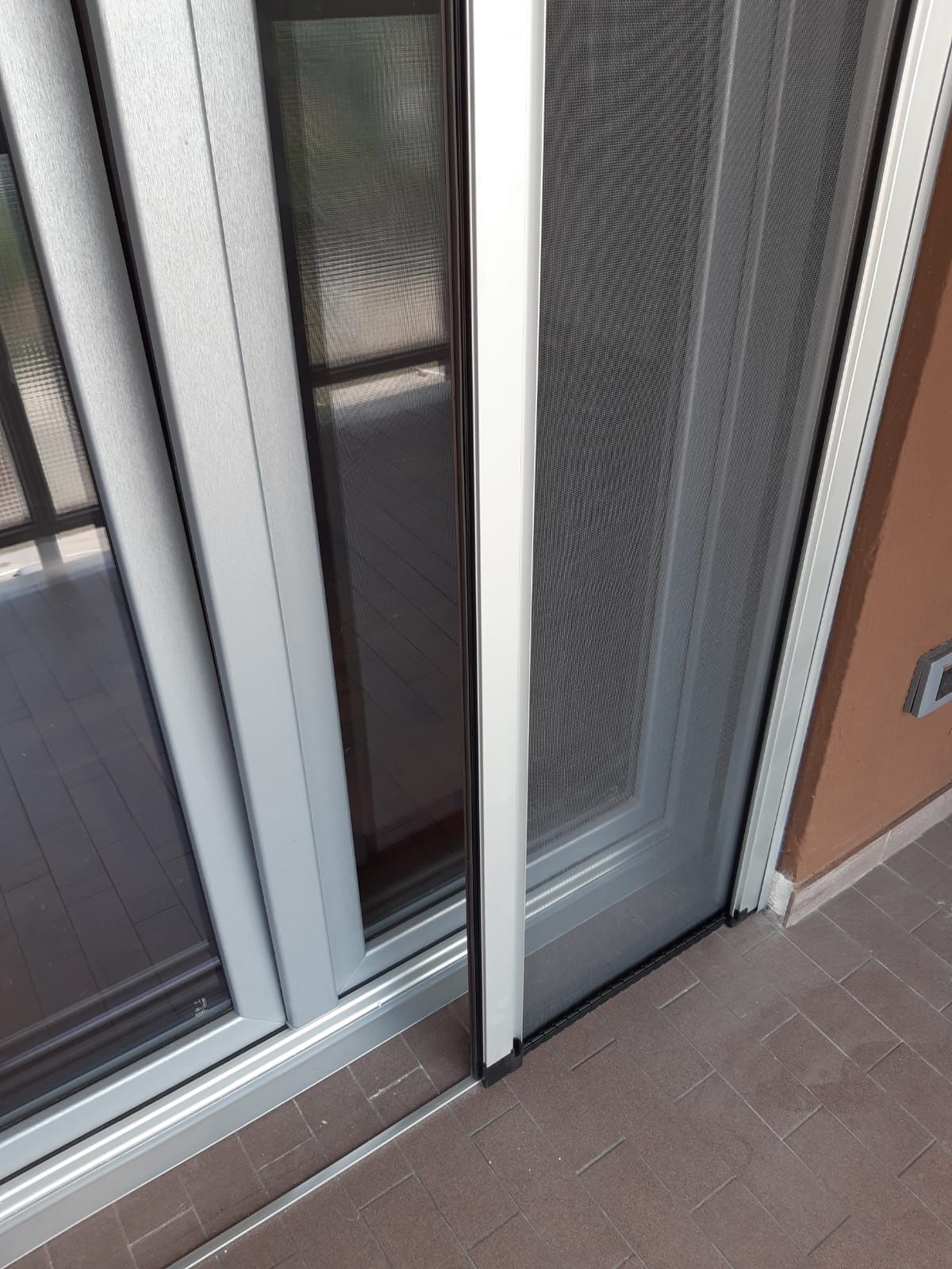 Primo piano di una porta a zanzariera con telaio bianco, parzialmente aperta, accanto a una porta a vetri su un patio in mattoni.
