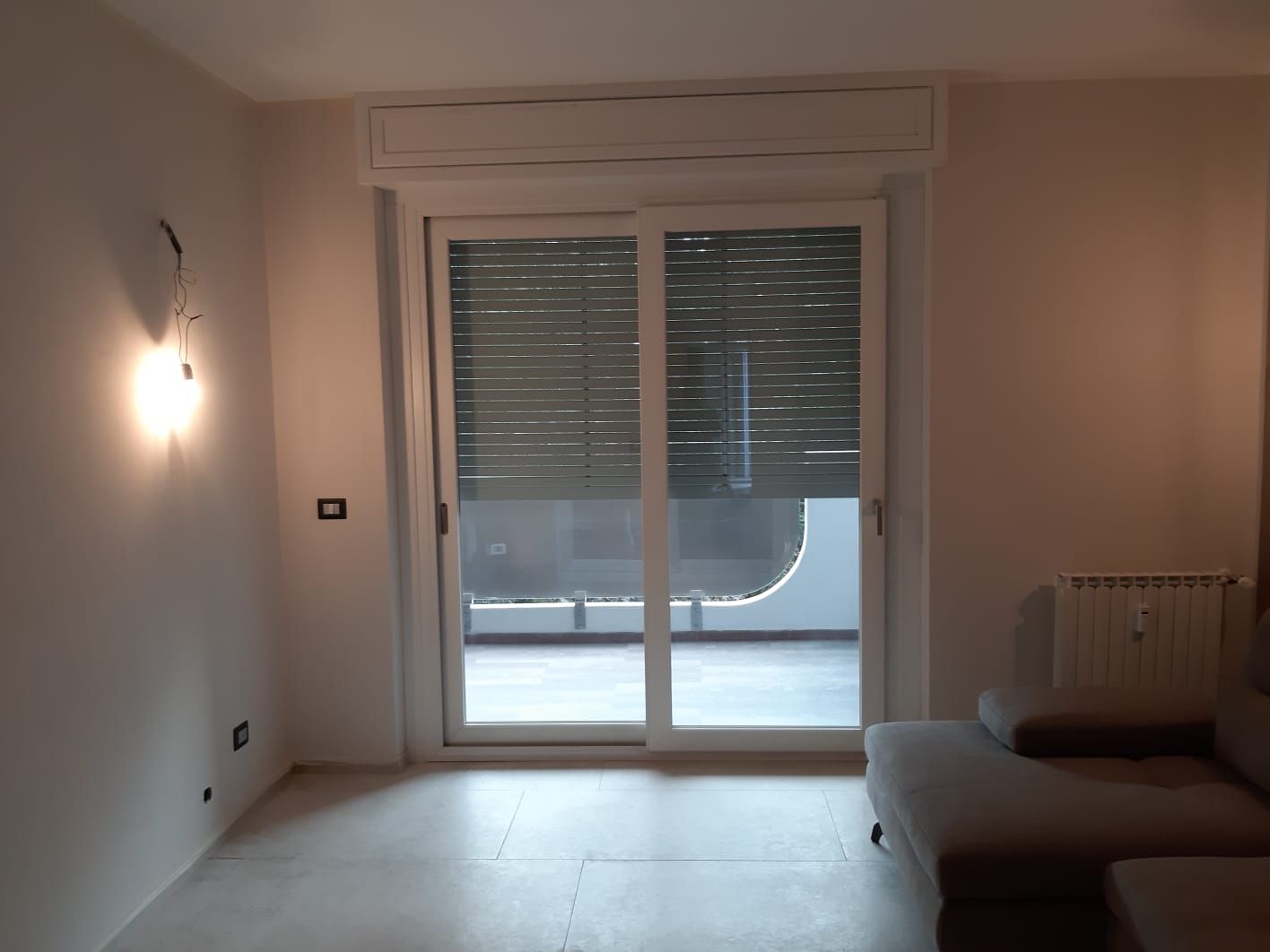 Un soggiorno con una porta scorrevole in vetro che si apre su un balcone, persiane chiuse. Pareti grigio chiaro, un divano e un'applique.