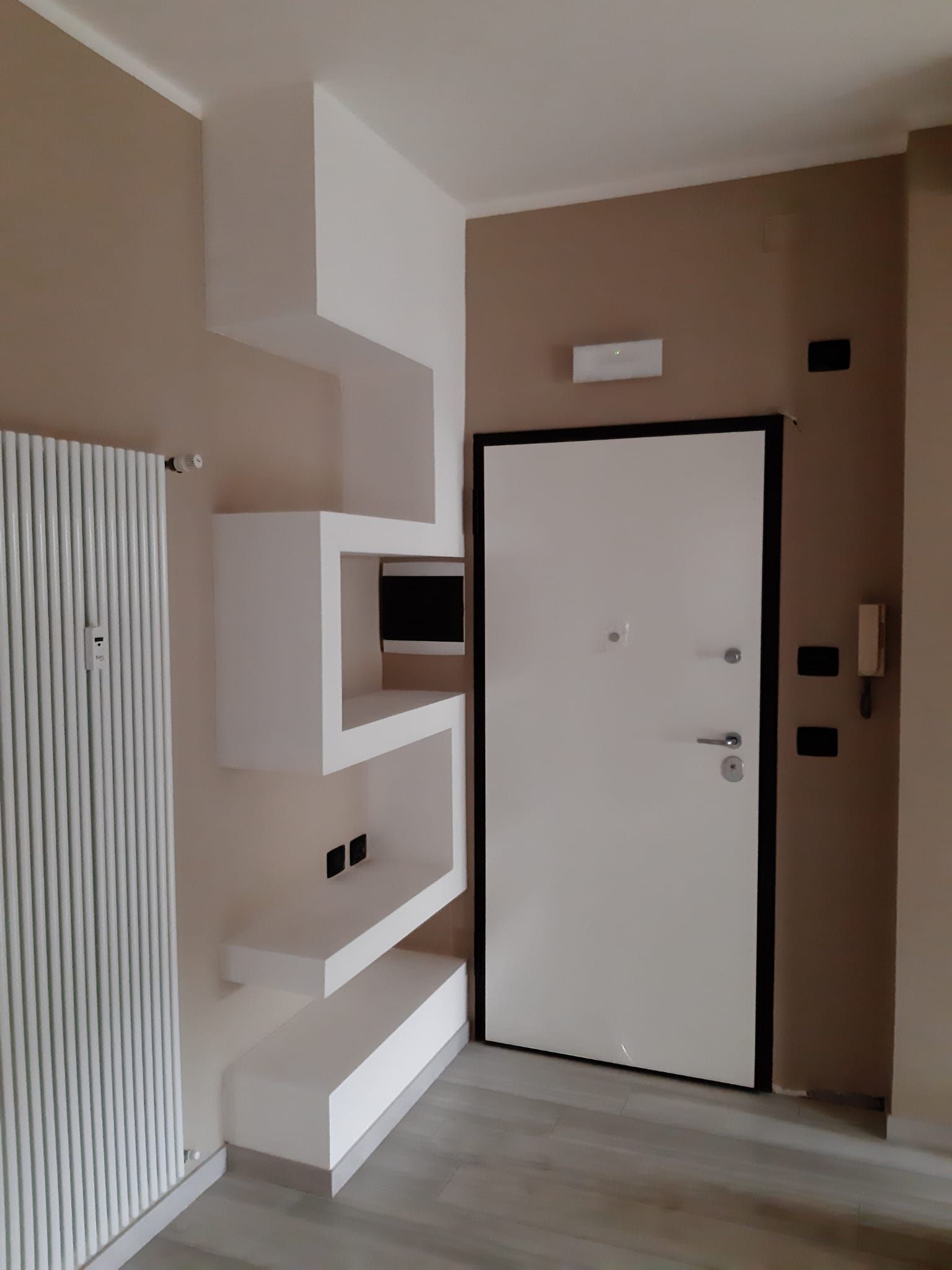 Ingresso con mensole bianche, una porta bianca e un termosifone verticale su una parete beige.