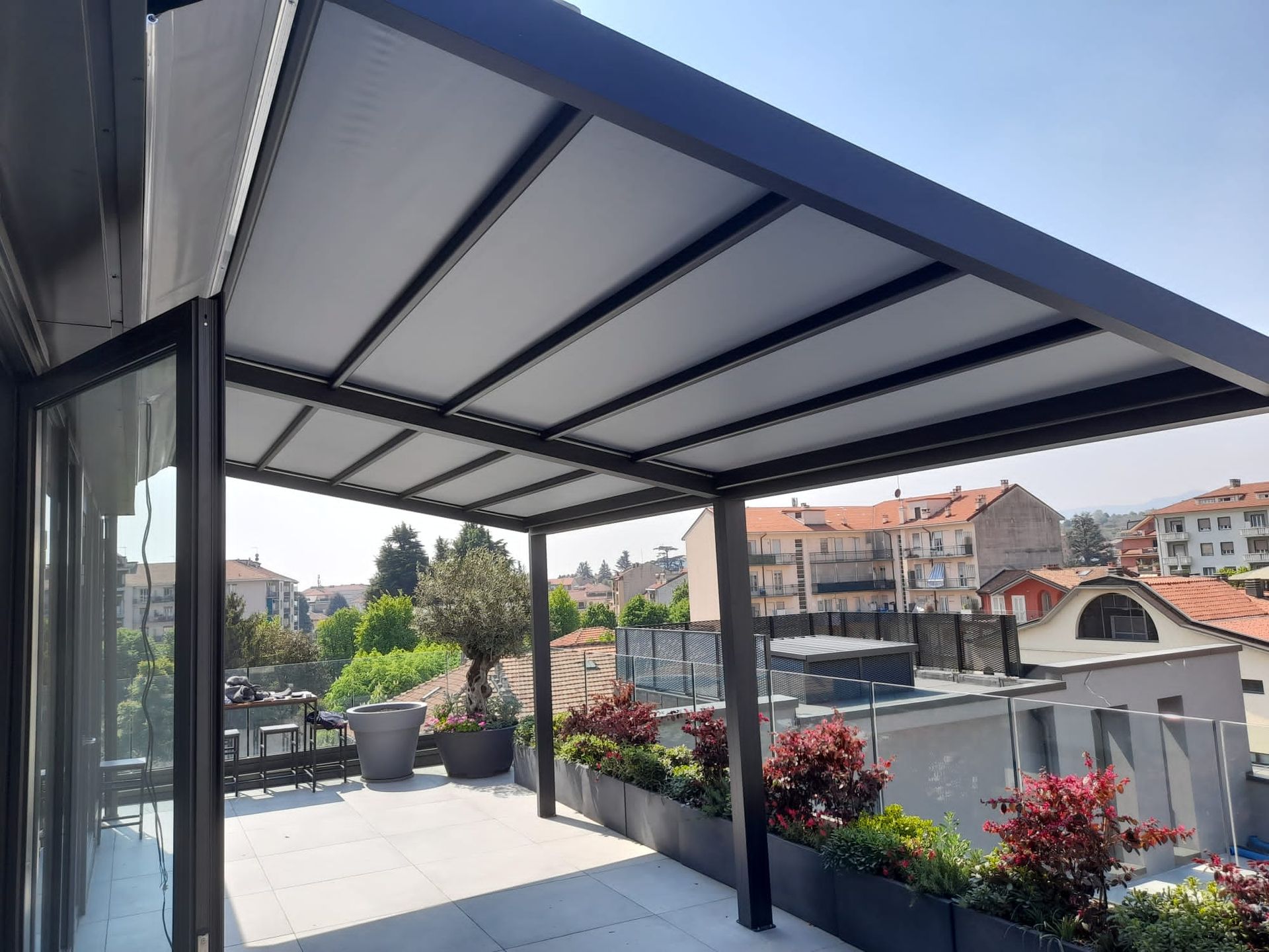 Pergola nera su un patio sul tetto, che fornisce ombra alle sedute ed è circondata da fioriere con piante rosse.