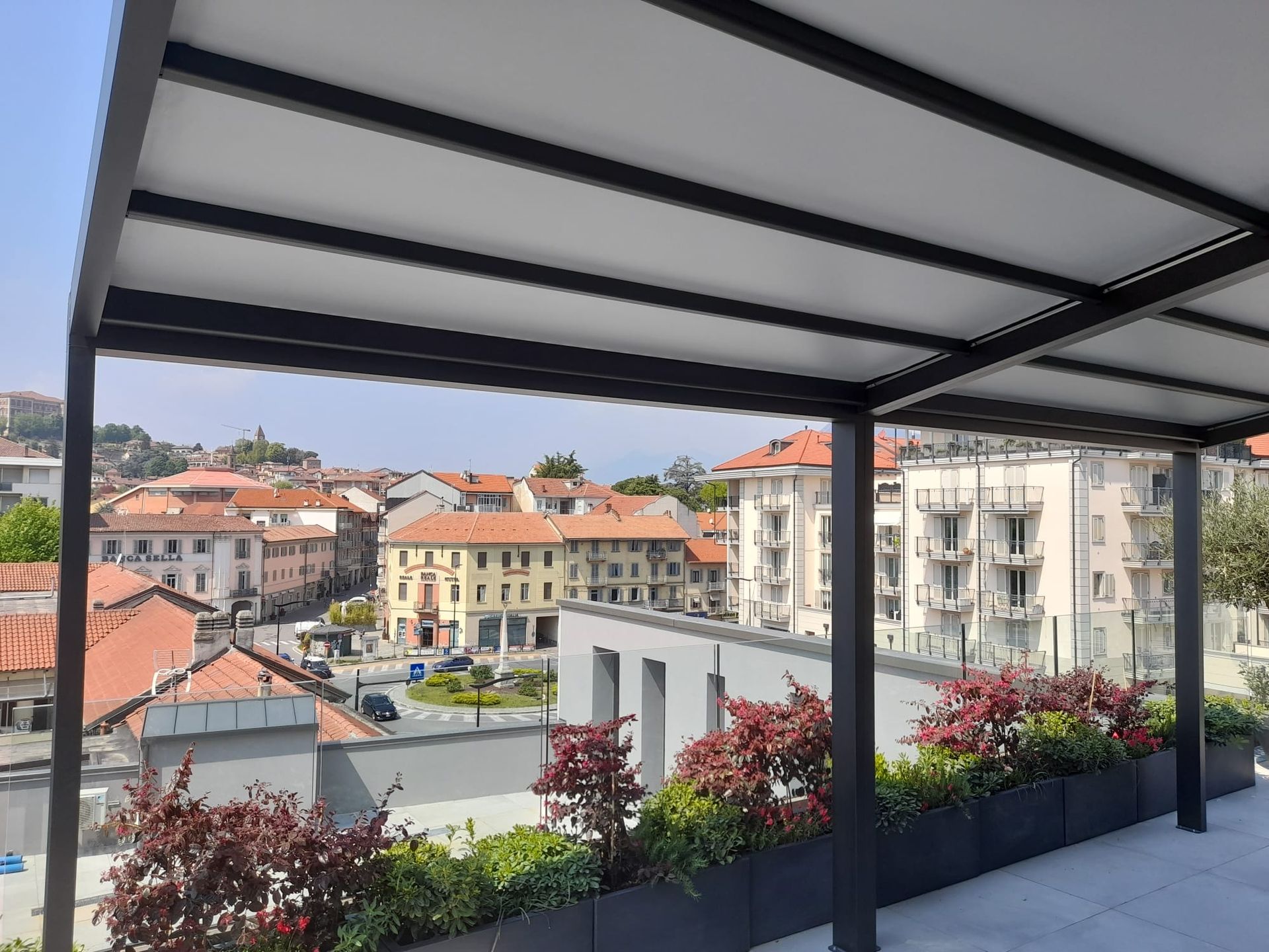 Pergola sul tetto con vista su una città europea. Struttura nera, tettoia bianca, edifici con tetti rossi.