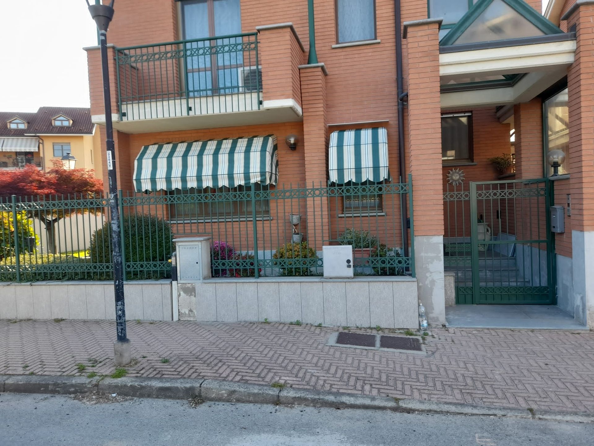 Casa in mattoni con tende da sole a strisce verdi e bianche, cancello in metallo e marciapiede.