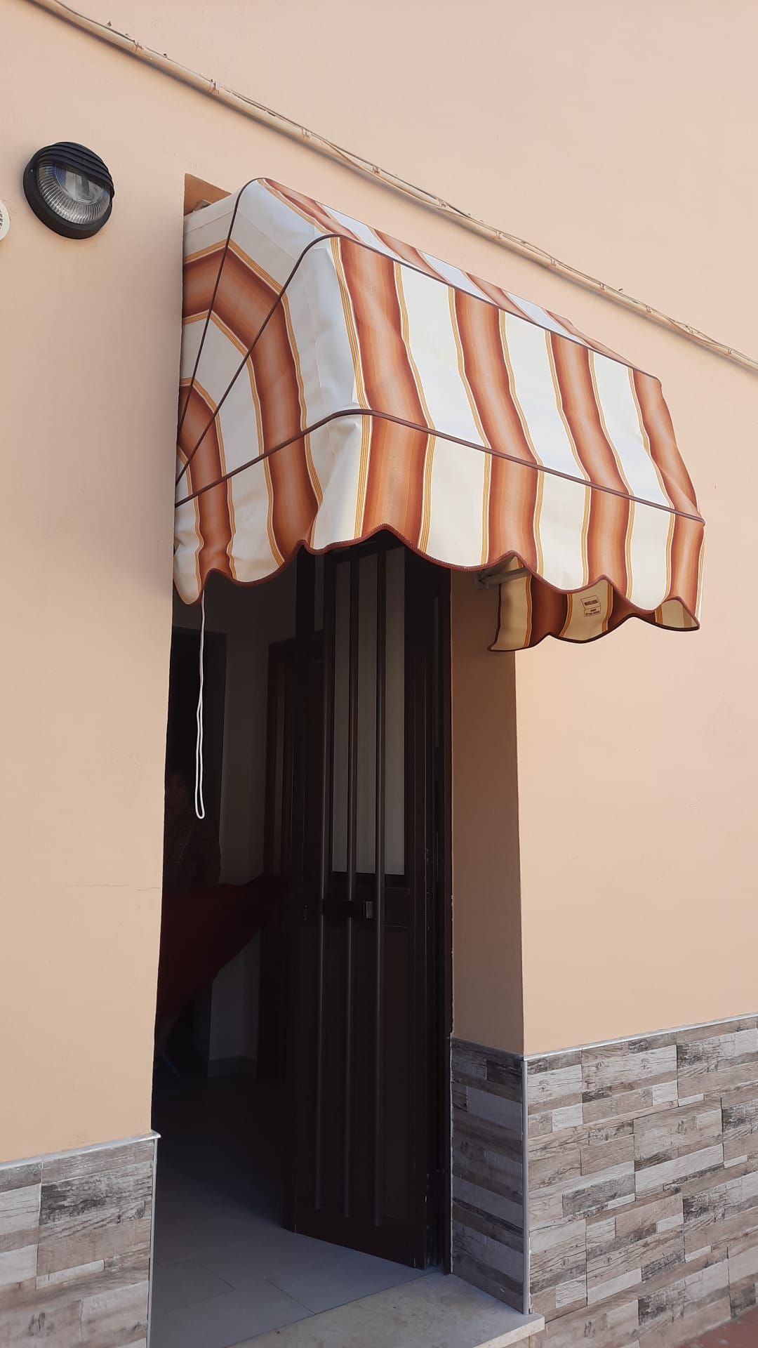 Porta con tenda a strisce, beige e marroni, sopra una porta di sicurezza in metallo di un edificio color pesca.