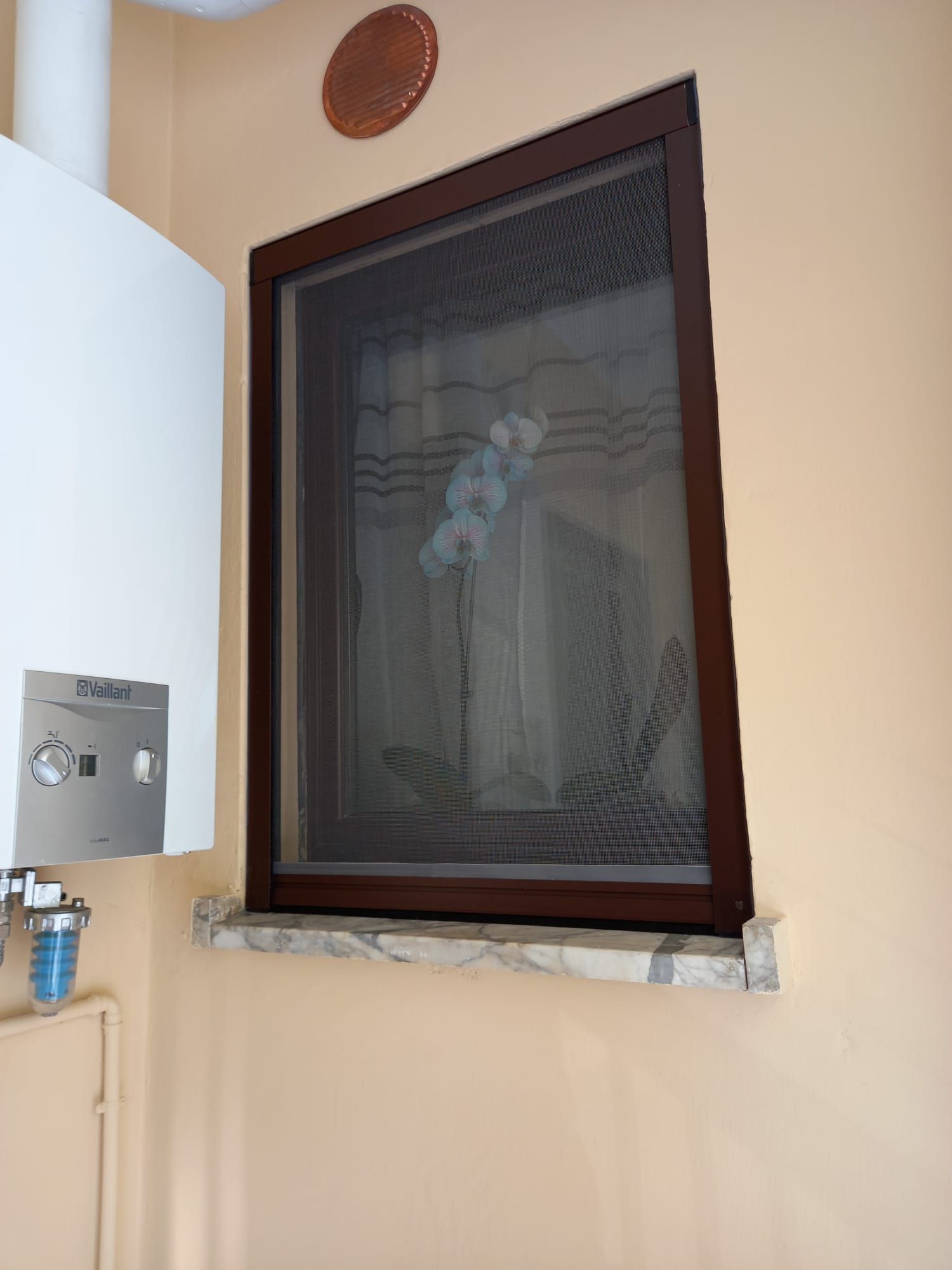Finestra con cornice marrone e zanzariera, montata su un davanzale in marmo. Uno scaldabagno è sulla sinistra. Pareti beige chiaro.