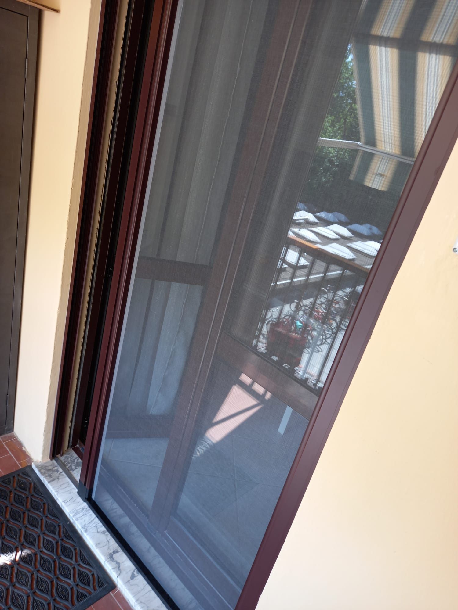 Porta scorrevole a zanzariera, rete scura, con telaio marrone. Pareti beige, zerbino scuro e balcone esterno riflesso.