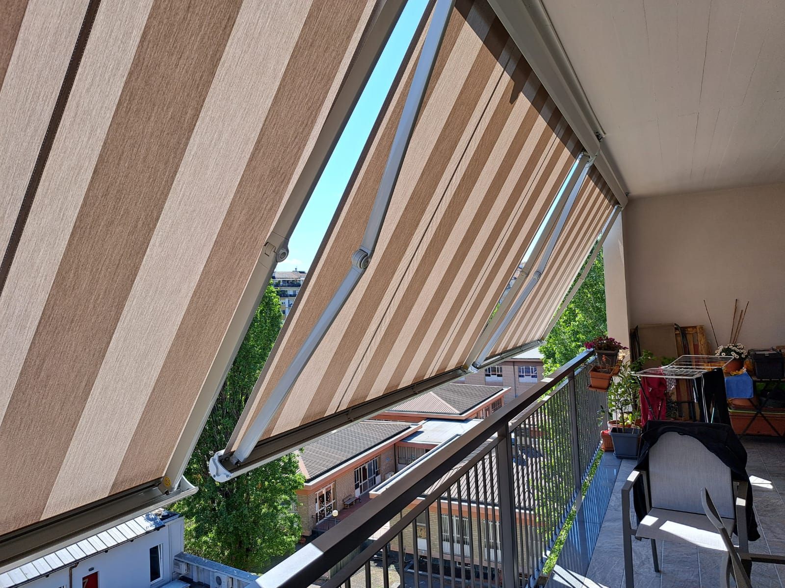 Tenda a strisce che si estendeva su un balcone, creando ombra con edifici e alberi sullo sfondo.