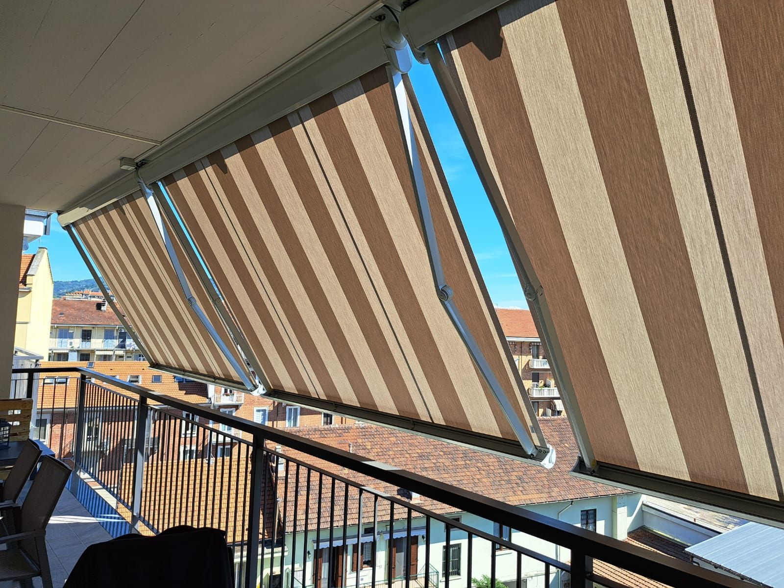 Balcone con tende da sole a strisce marroni e beige estese per creare ombra. Vista sulla città con cielo azzurro.