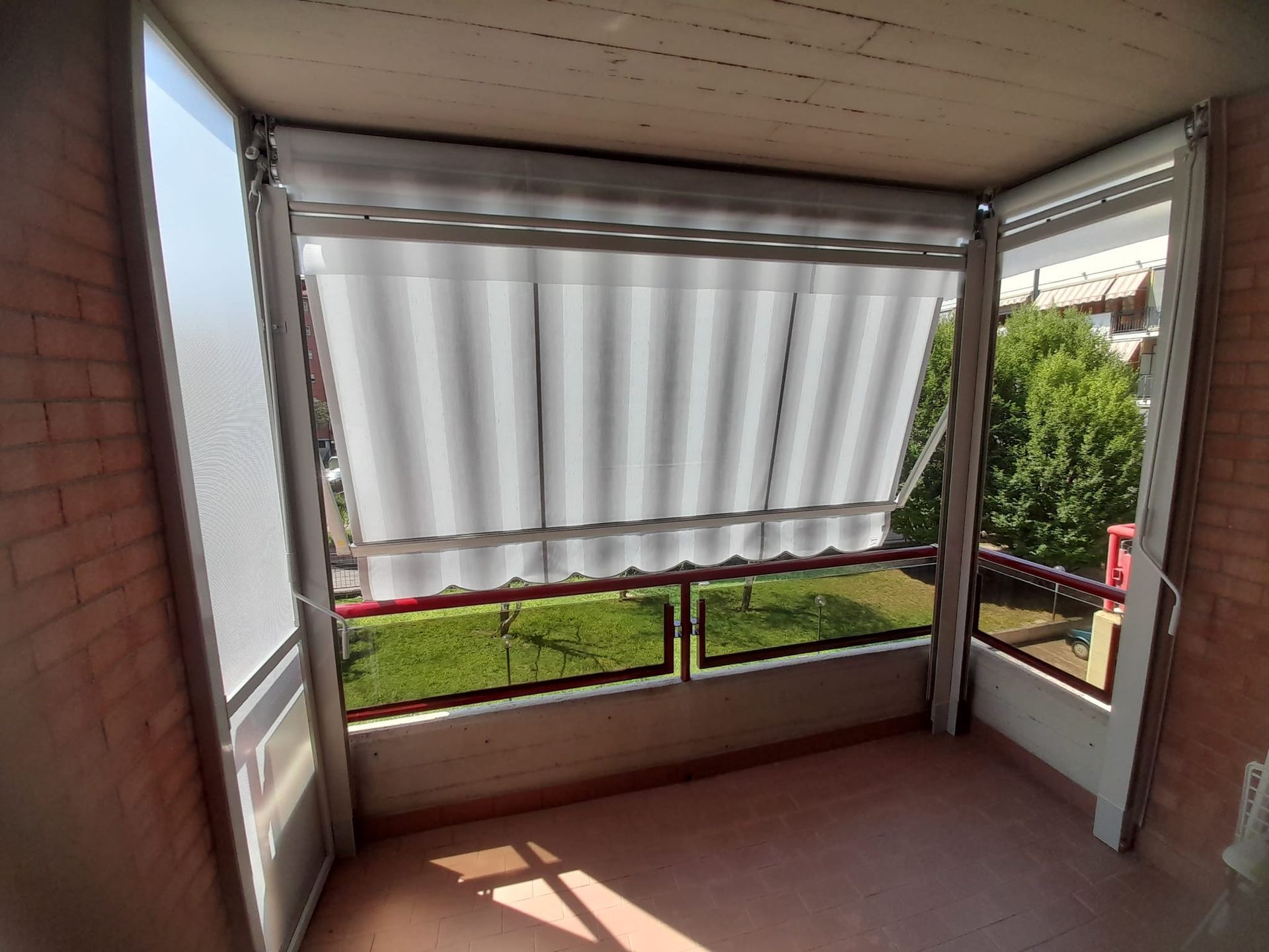 Un balcone con una tenda bianca, ringhiere rosse e vista sull'erba verde e sugli alberi.