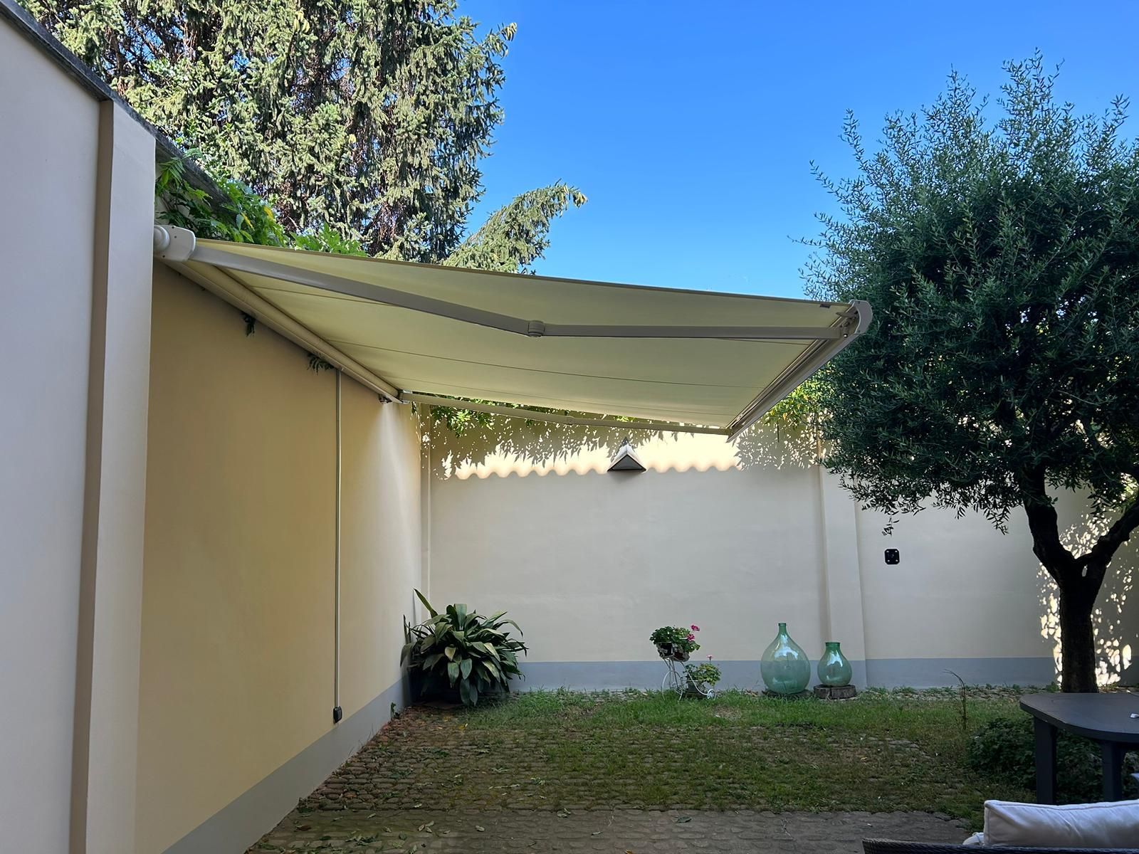 Tenda da sole beige che ombreggia un patio nel cortile sul retro con prato verde e piante contro un muro beige.