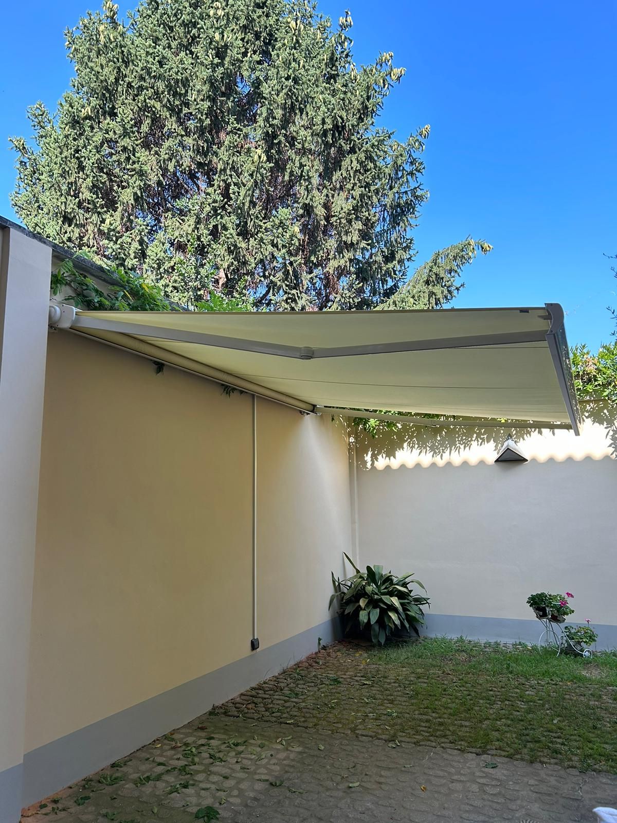 Una tenda da sole retrattile color crema su una parete beige, sopra una piccola area erbosa con piante e un cielo azzurro.