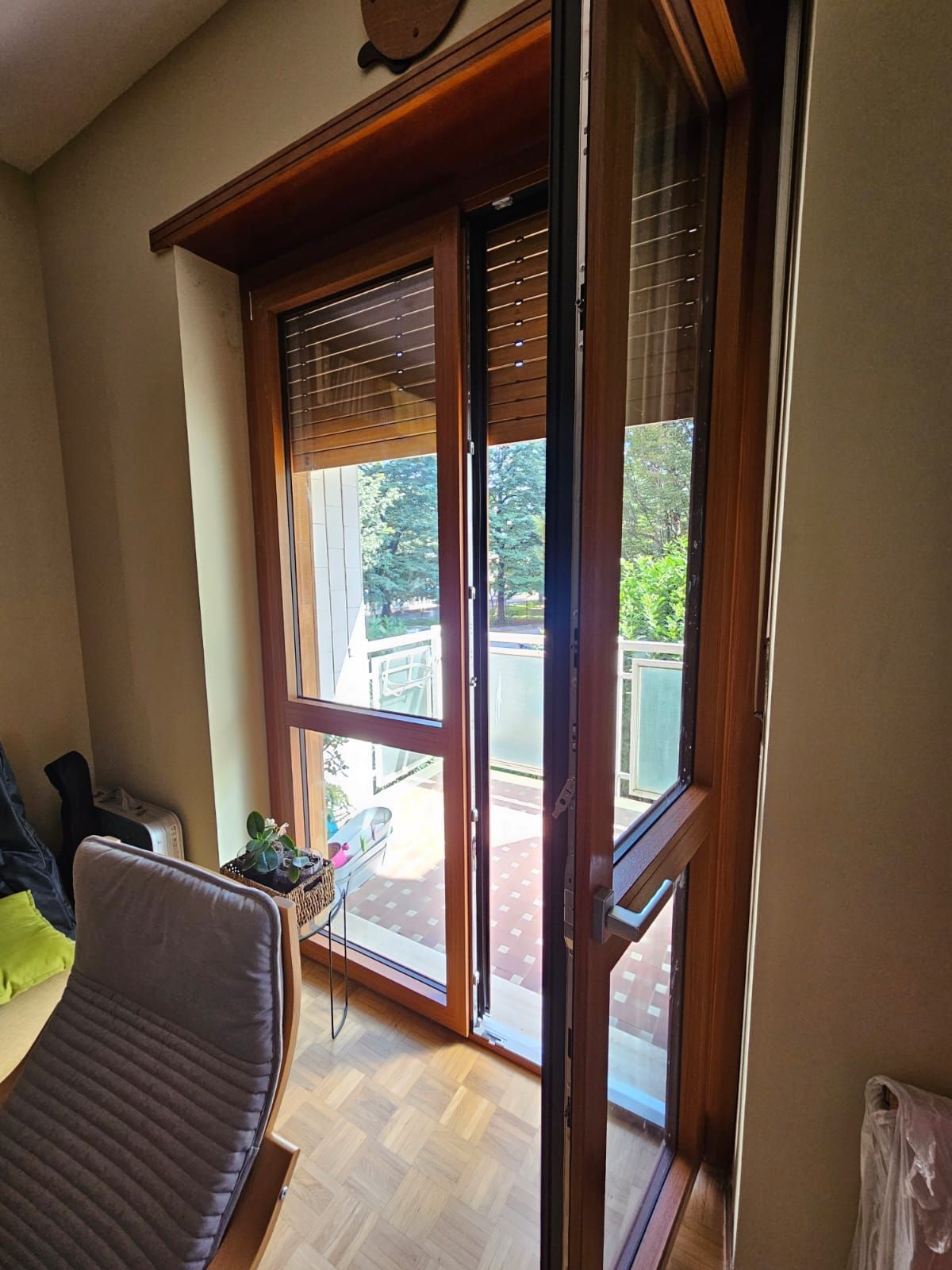 Porta a vetri con cornice in legno che si apre su un balcone, con una sedia seduta in primo piano e alberi verdi all'esterno.