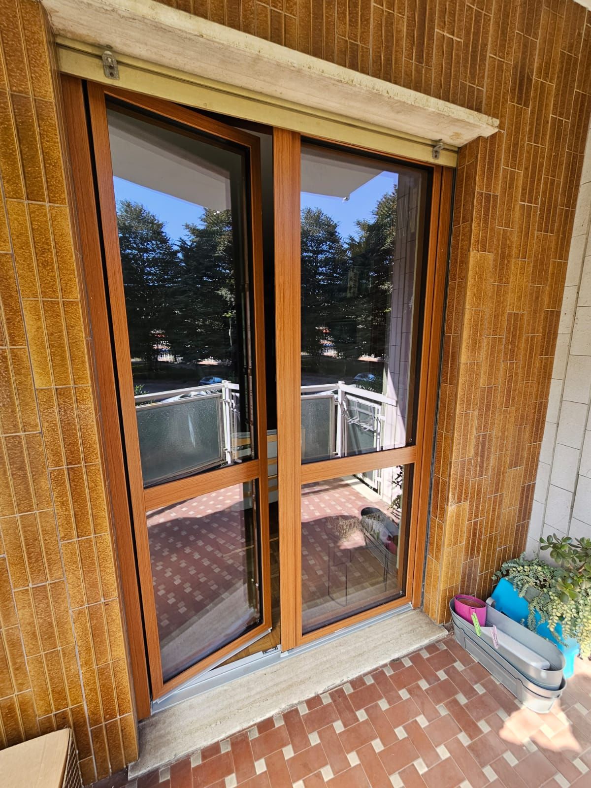 Porte a vetri con cornice marrone su un balcone. Pavimento in mattoni, rivestimento murale intrecciato e scorcio di alberi e mobili.
