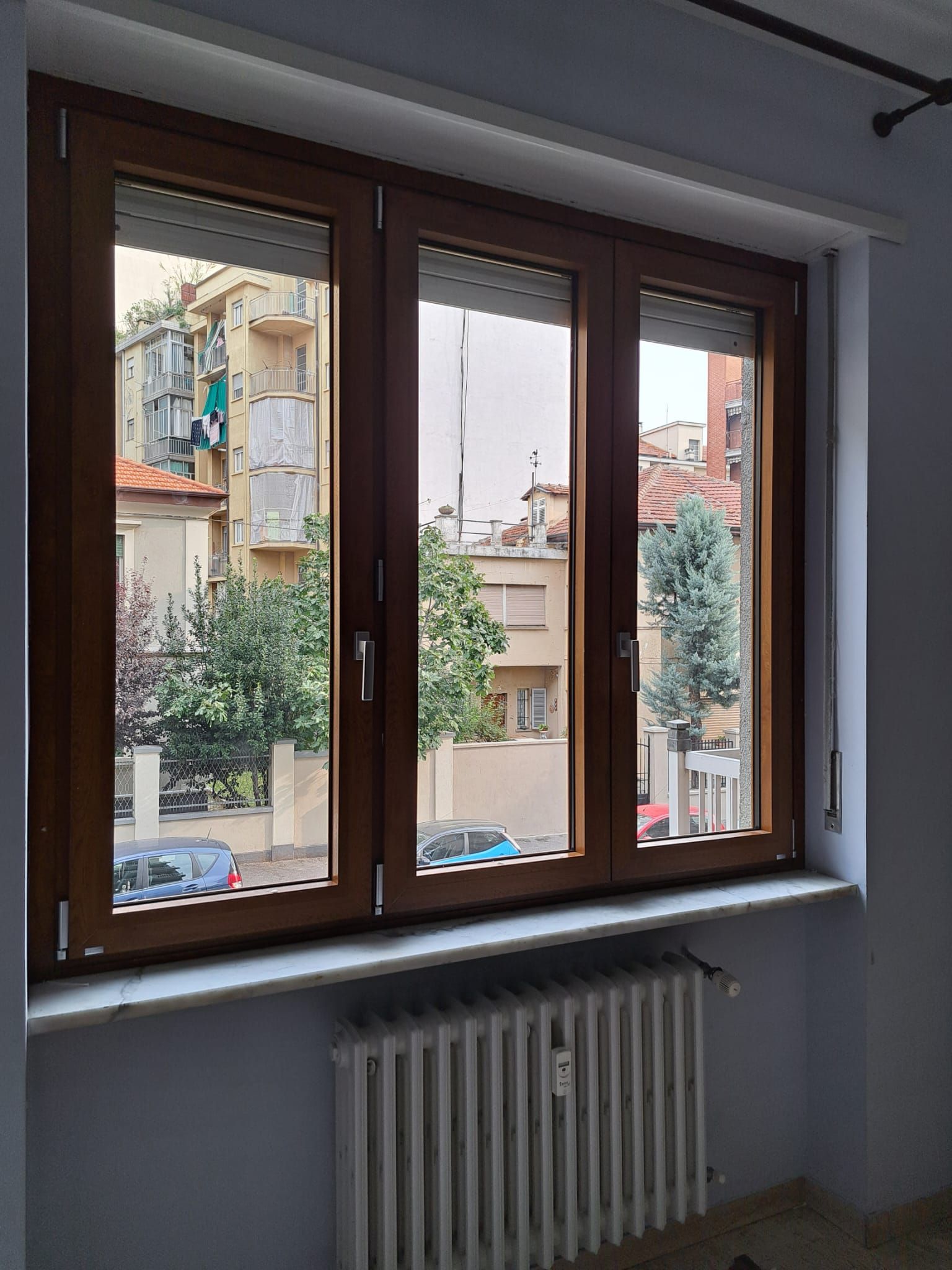 Finestra con cornice marrone che si affaccia su un panorama cittadino con edifici, alberi e automobili. Sotto la finestra c'è un termosifone.