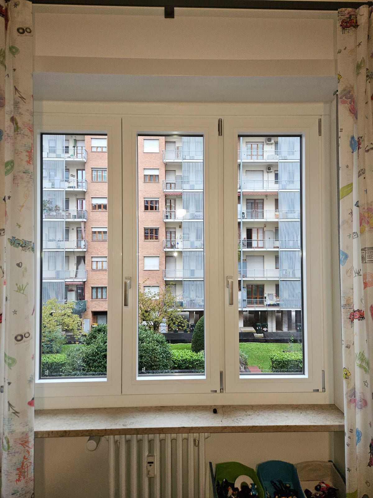 Una finestra a tre ante che si affaccia su un condominio e su un cortile. Cornice bianca, tende su entrambi i lati.