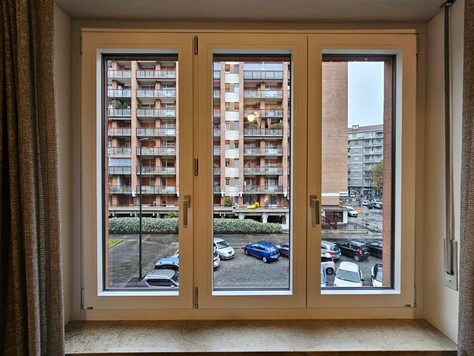 Vista da tre finestre di condomini e auto parcheggiate in una giornata nuvolosa.