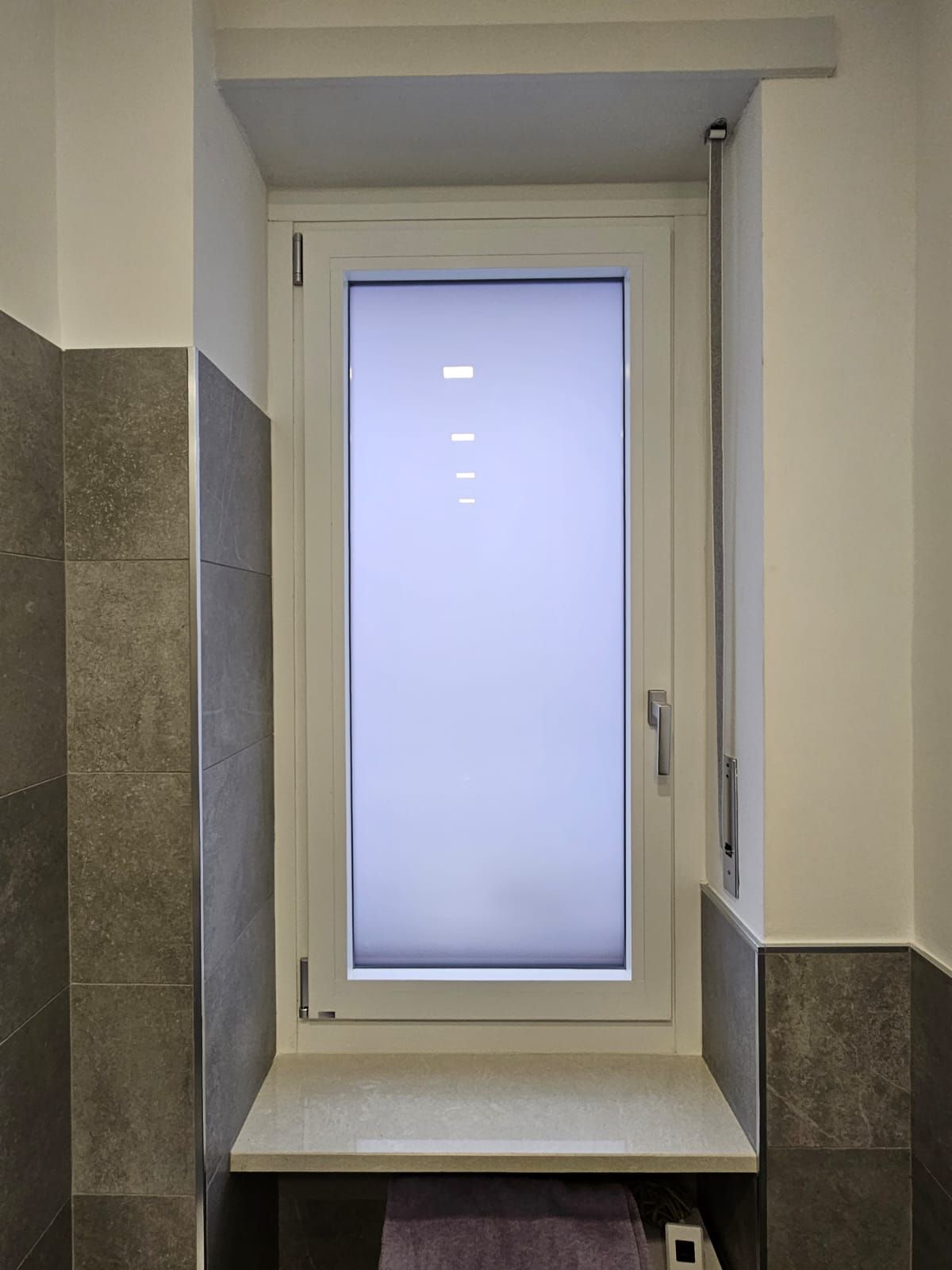 Una finestra con cornice bianca e vetro smerigliato in un bagno. Piastrelle grigie su entrambe le pareti.