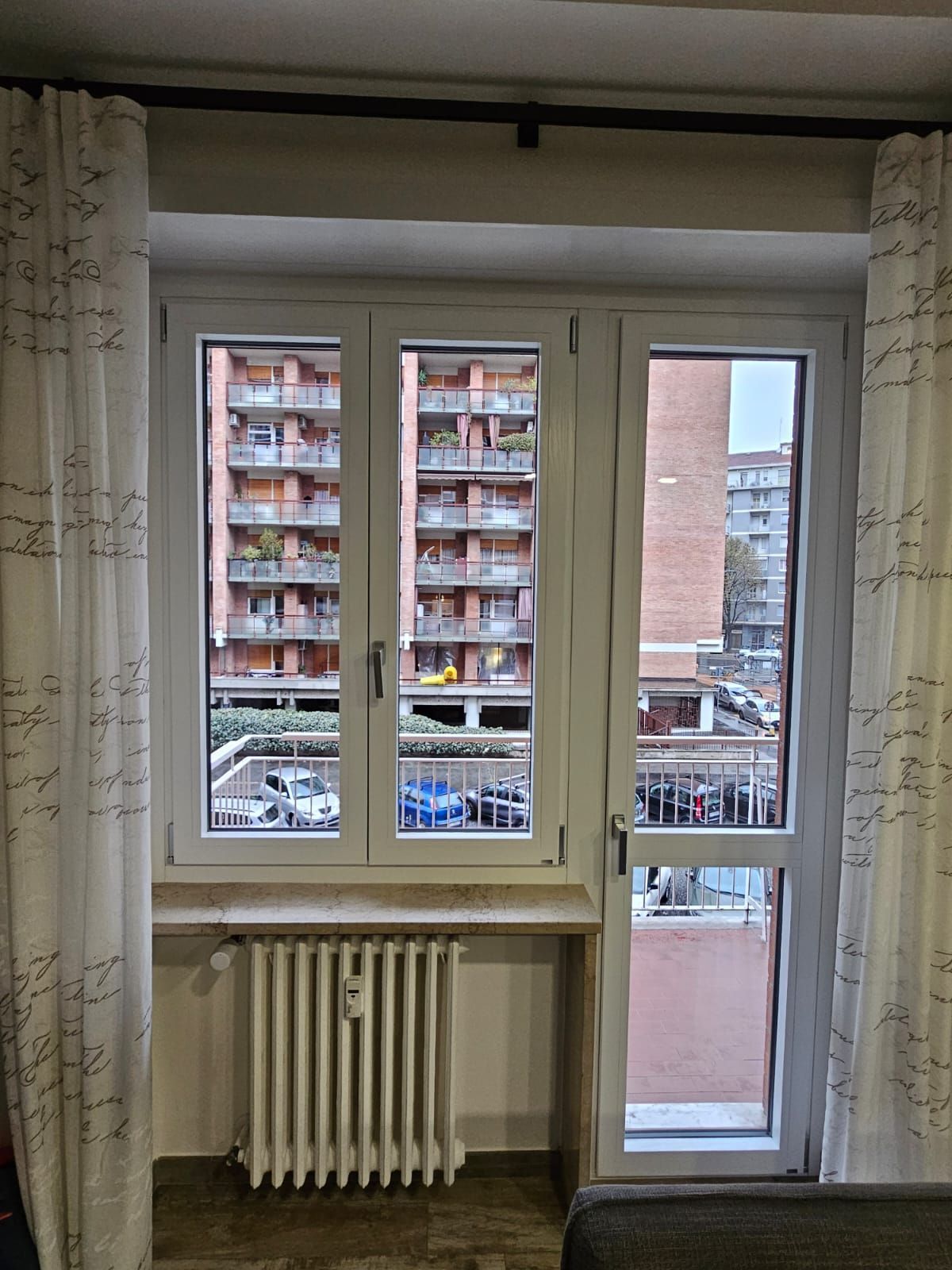 Vista dalla finestra: finestra con cornice bianca e tende beige, che mostra un condominio e delle auto parcheggiate.