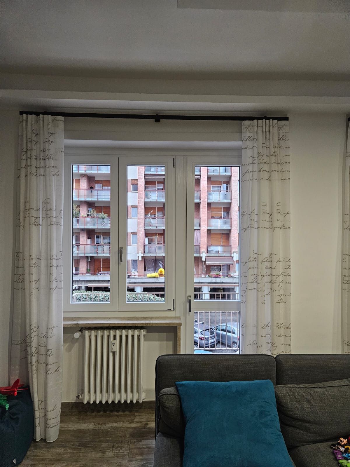 Finestra con tende bianche trasparenti, termosifone, vista sull'edificio, divano con cuscino verde acqua.